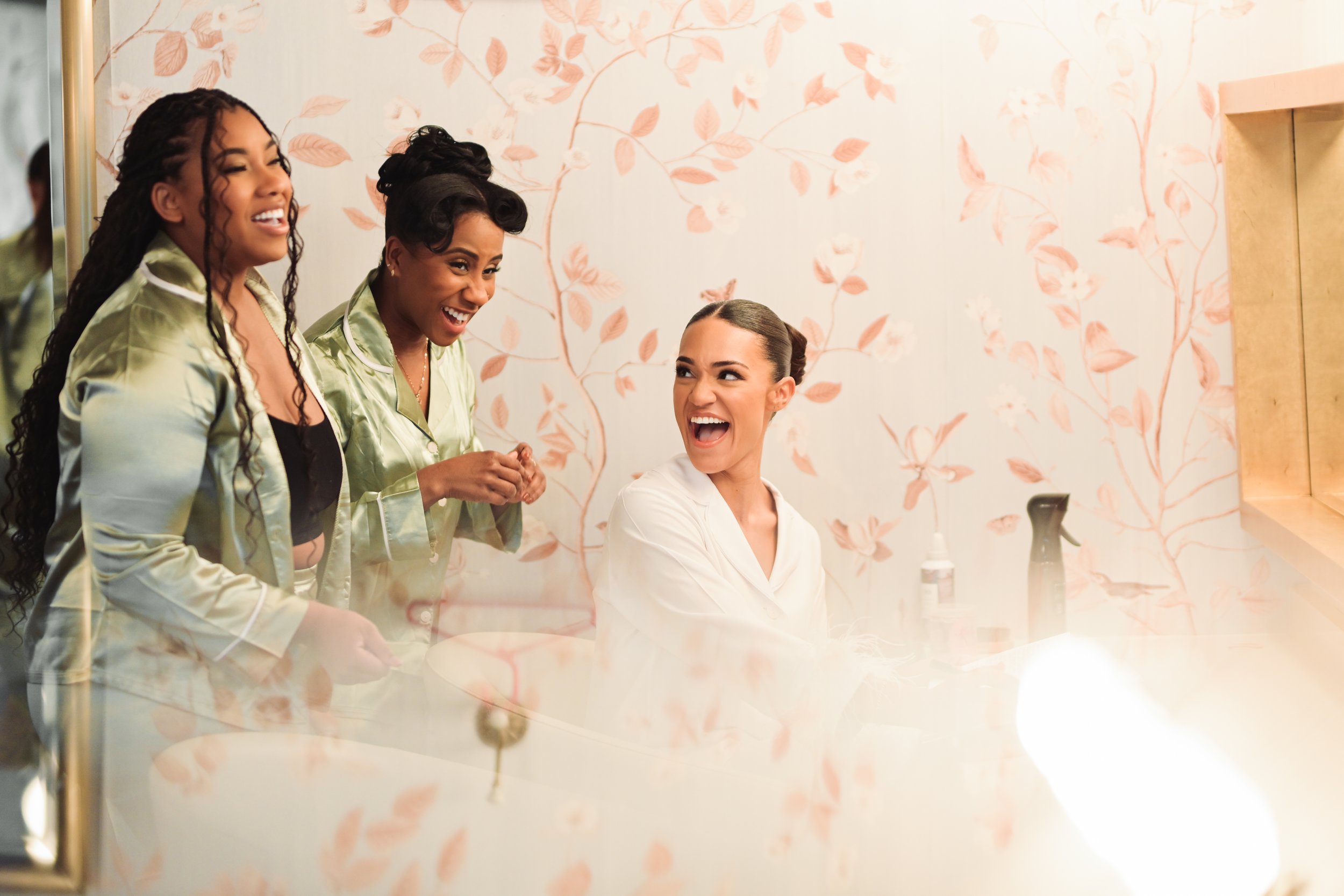 Three women in luxury bridal suite laughing and talking in front of a mirror with a pink leaf pattern on the wall.