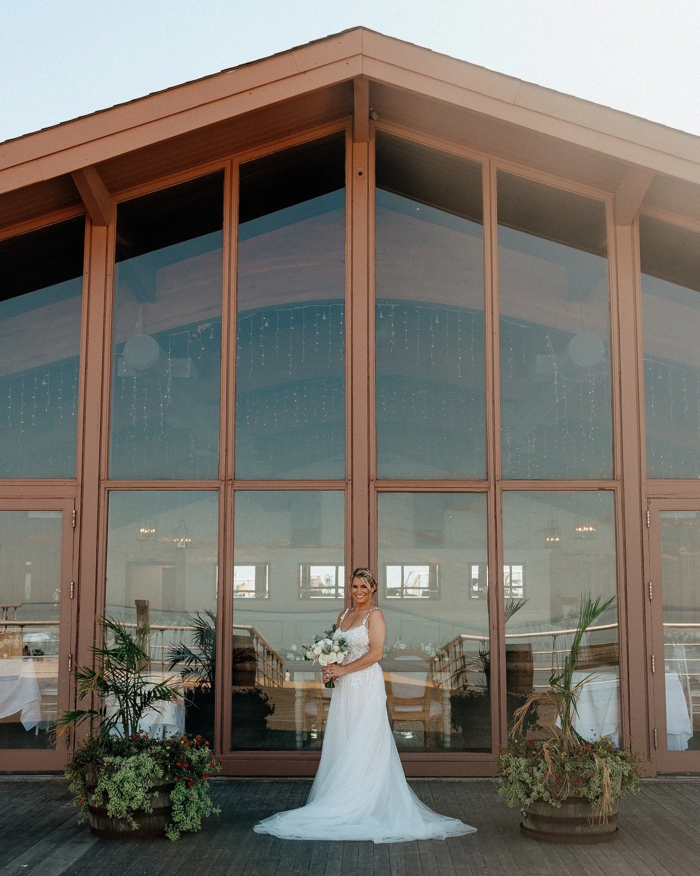 A woman in a white wedding dress holding a bouquet of white flowers, standing in front of a large glass window with plants and fairy lights inside.