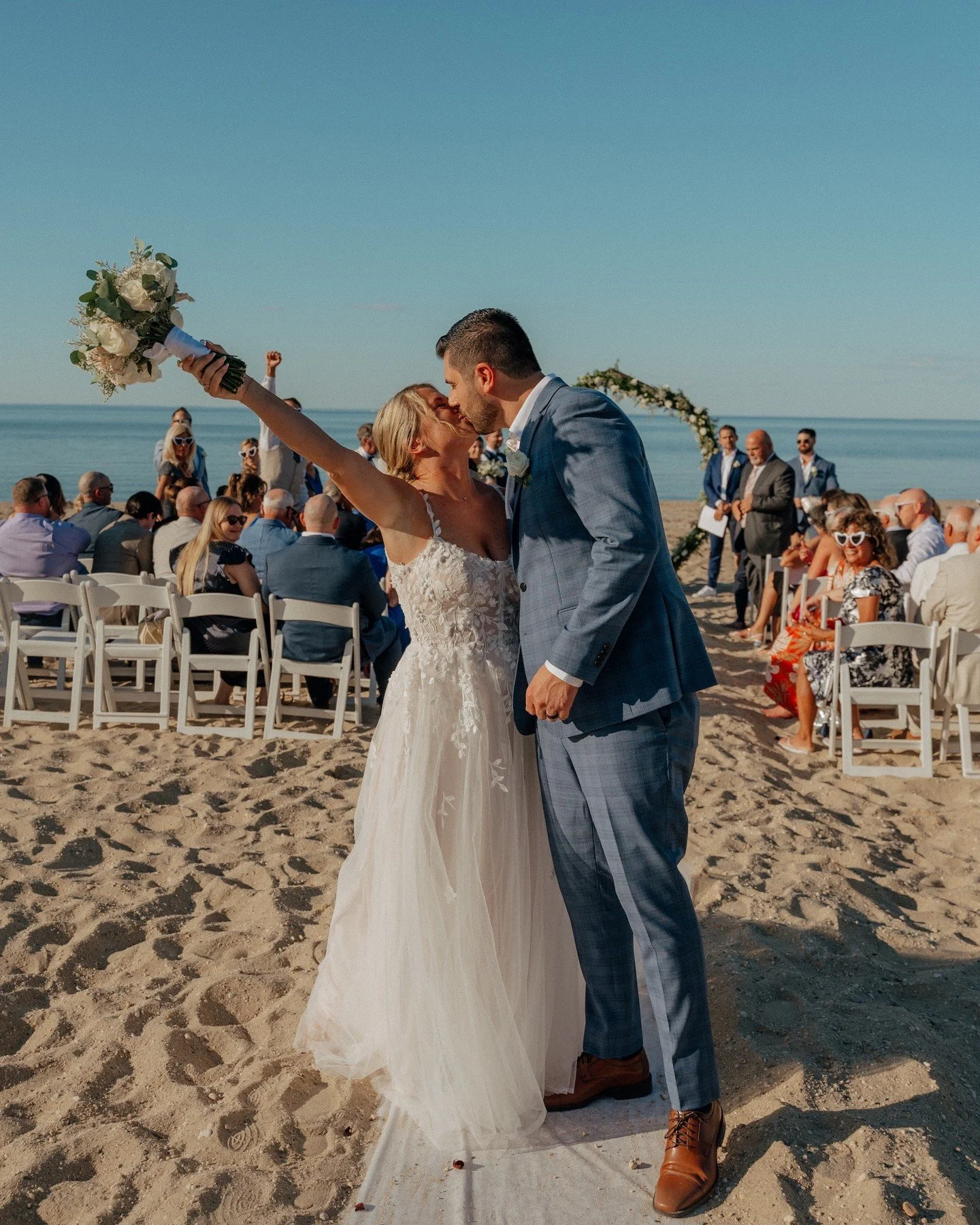 A bride and groom kiss during their beach wedding. The bride holds a bouquet in the air, while guests sit and stand in the background.