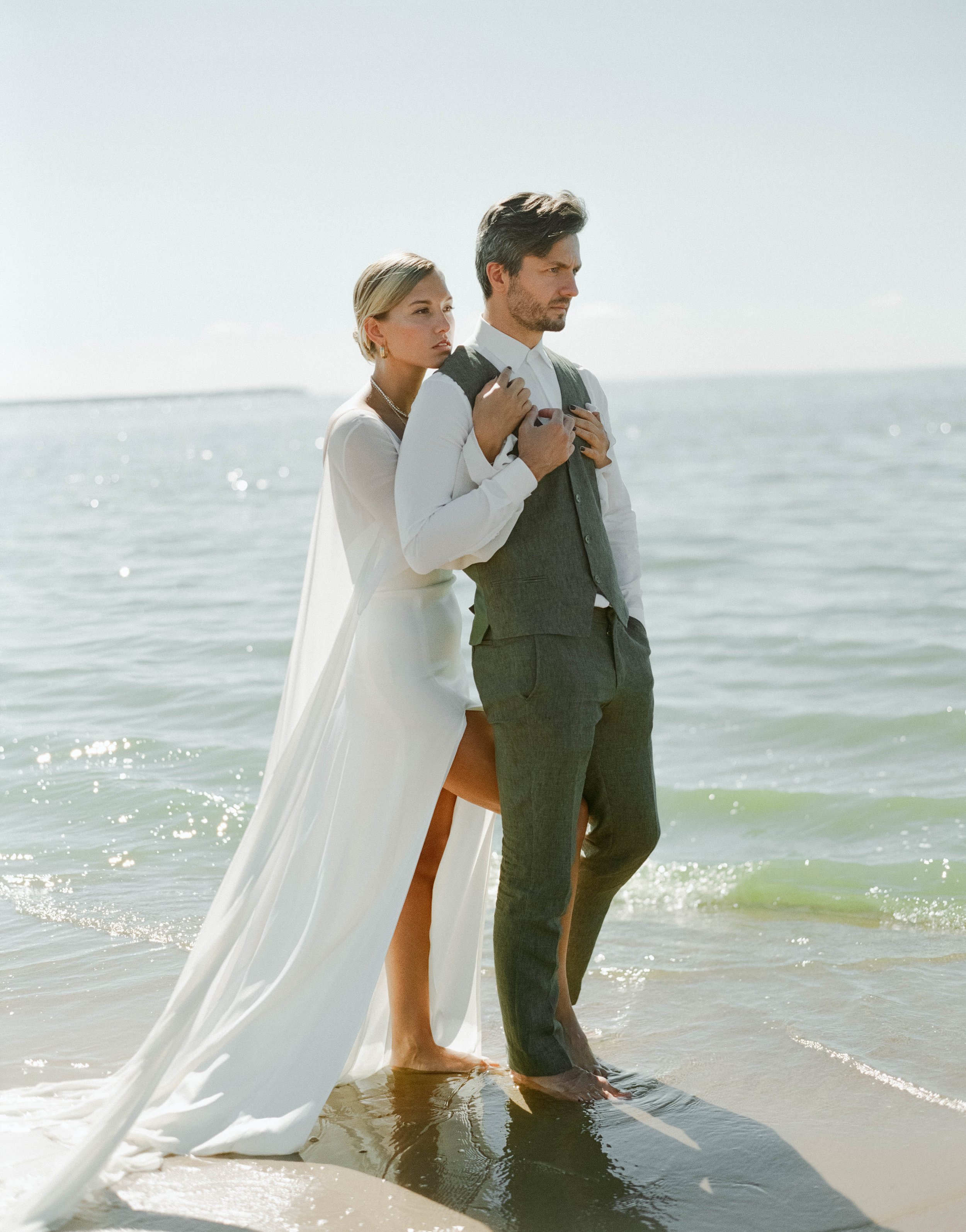 A man and woman standing in shallow ocean water, facing the horizon with serious expressions. The woman is holding the man's shoulder, dressed in a white dress, and the man is wearing a white shirt with a gray vest and pants. The background features a calm sea and a light, cloudy sky.
