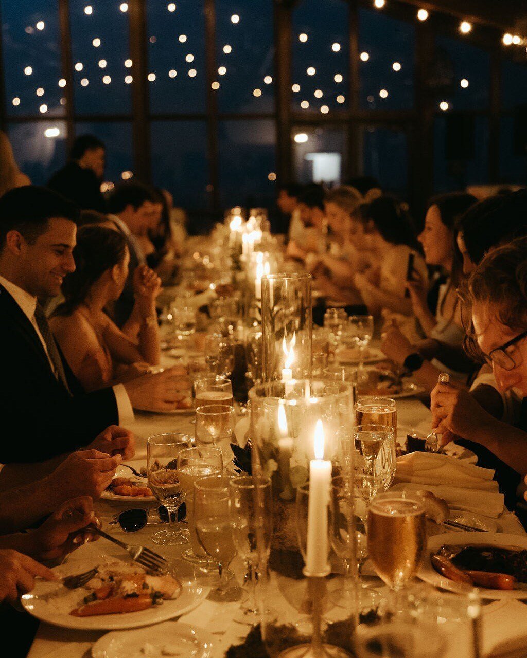 A formal dinner setting with a long table decorated with candles, where people are dressed in formal attire, enjoying a meal in a warmly lit, elegant venue with string lights overhead.