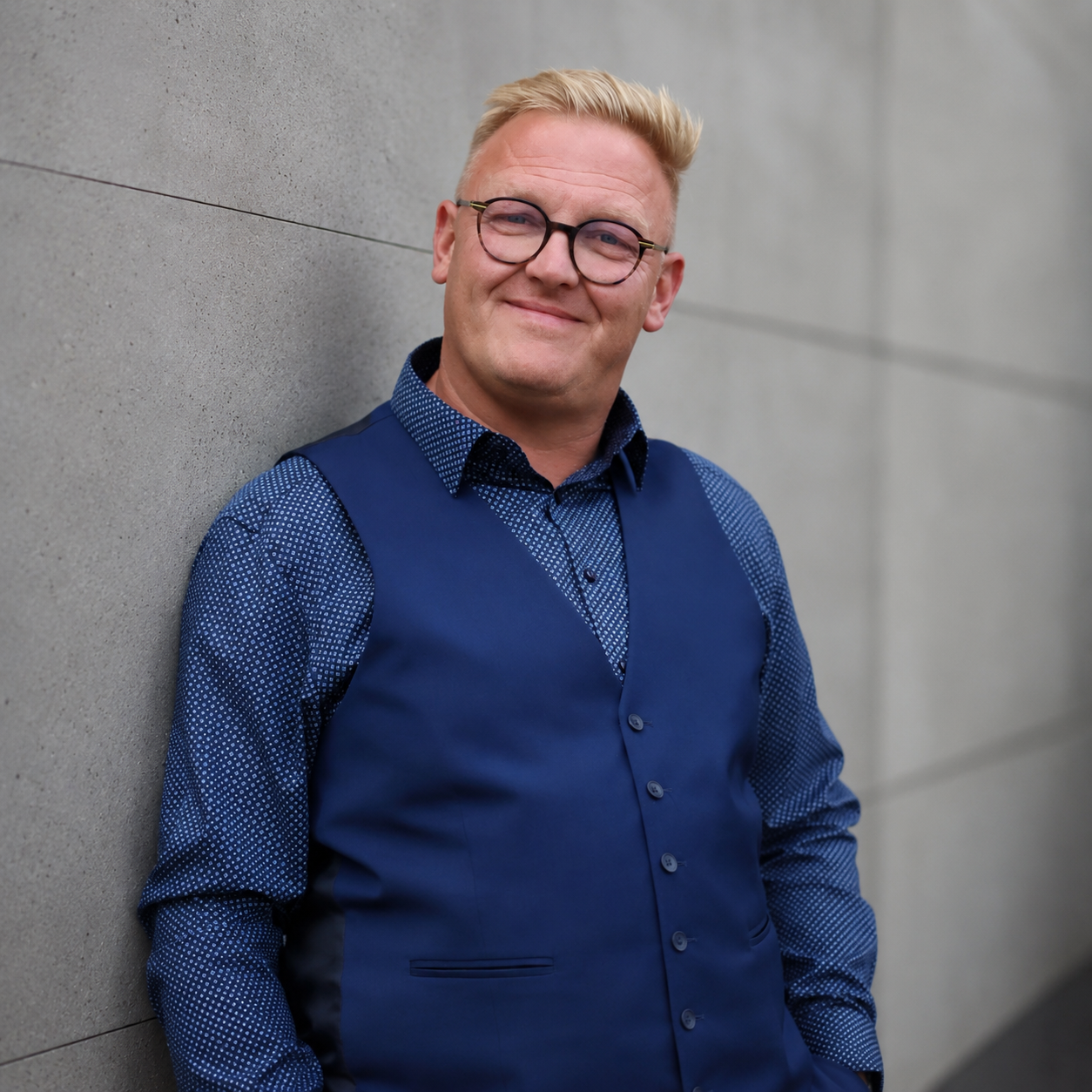 Ein lächelnder Mann mit Brille, blonder Kurzhaarschnitt, trägt eine blaue Weste und Hemd, lehnt an einer grauen Betonwand.