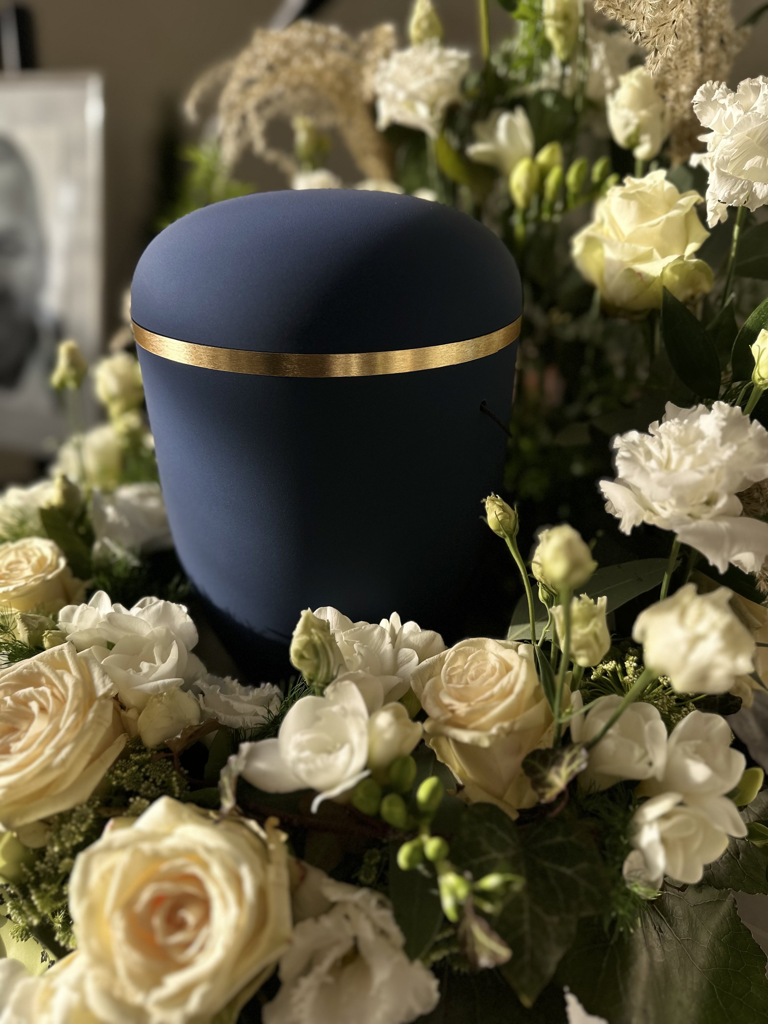 Ein blauer, runder Gegenstand mit einer goldfarbenen Linie umrahmt, liegt auf einem Blumenarrangement aus weißen und cremefarbenen Blumen, unter anderem Rosen, und grünem Blattwerk.