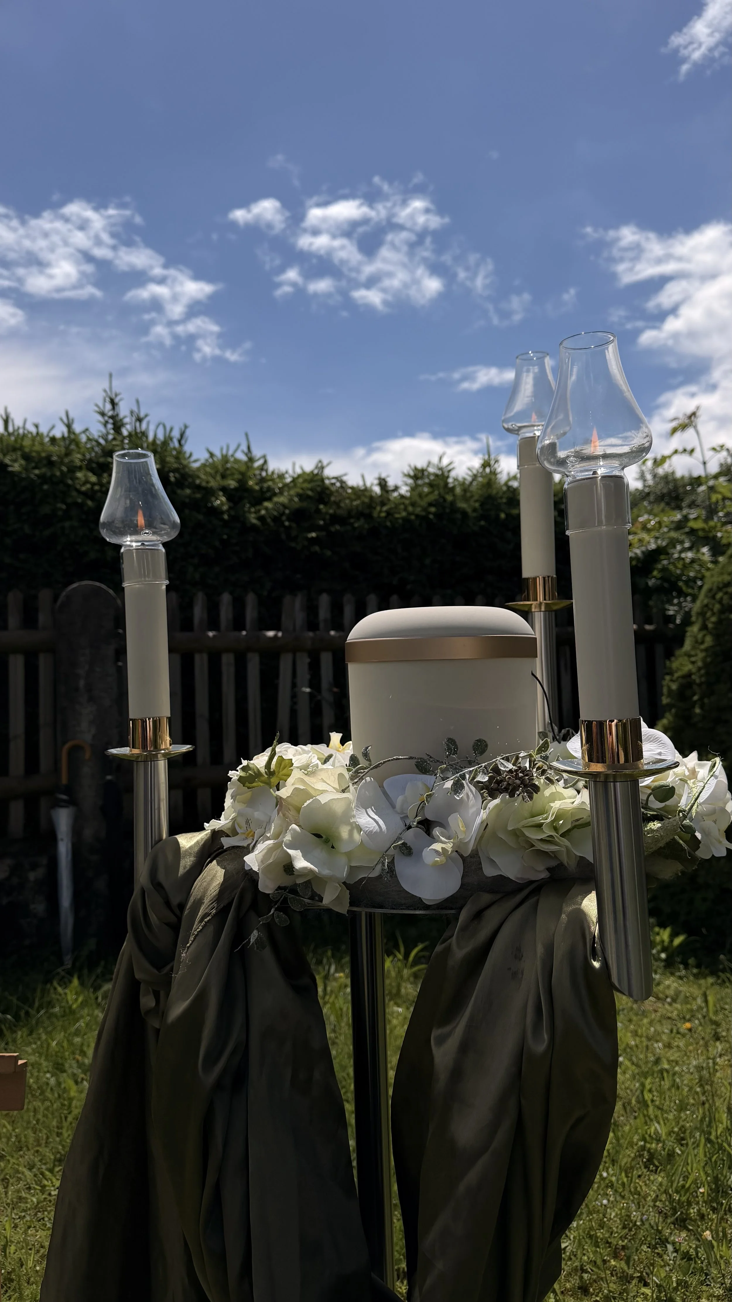 Ein Dekorationsstück im Freien mit weißen Blumen, Kerzenhaltern und einem weißen Behälter auf einer schwarzen Ständer, vor einem Holzzaun und einem blauen Himmel mit weißen Wolken.