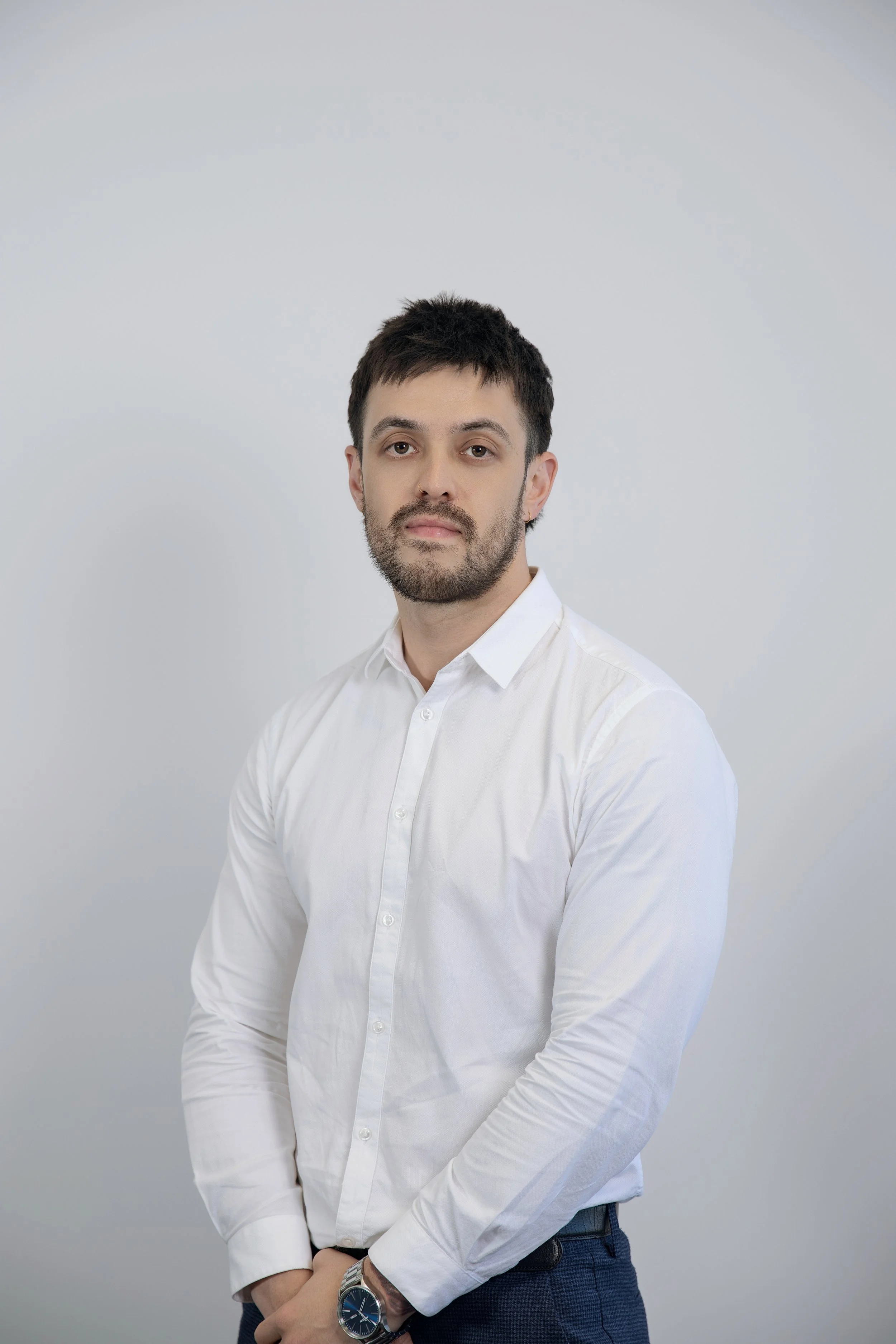 A young man with dark hair and a beard wearing a white dress shirt, dark pants, and a watch, standing against a light gray background. This is the principal mortgage broker of the company, he is wearing a blue a watch. He is smiling slightly