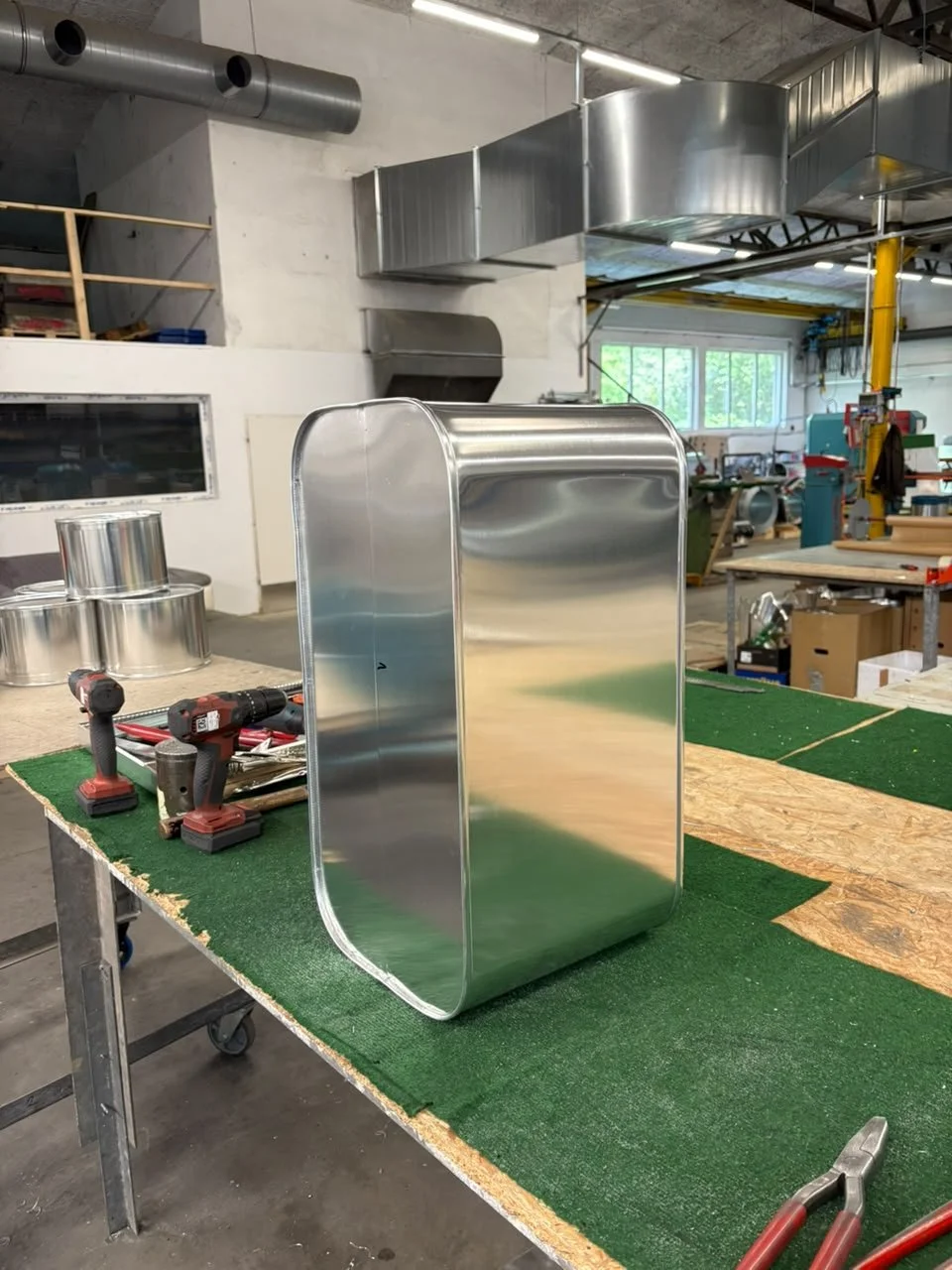 Ein Metallbehälter mit abgerundeten Ecken steht auf einem Werkstatttisch, umgeben von Elektrowerkzeugen und anderen Werkstücken in einer Fabrikhalle.