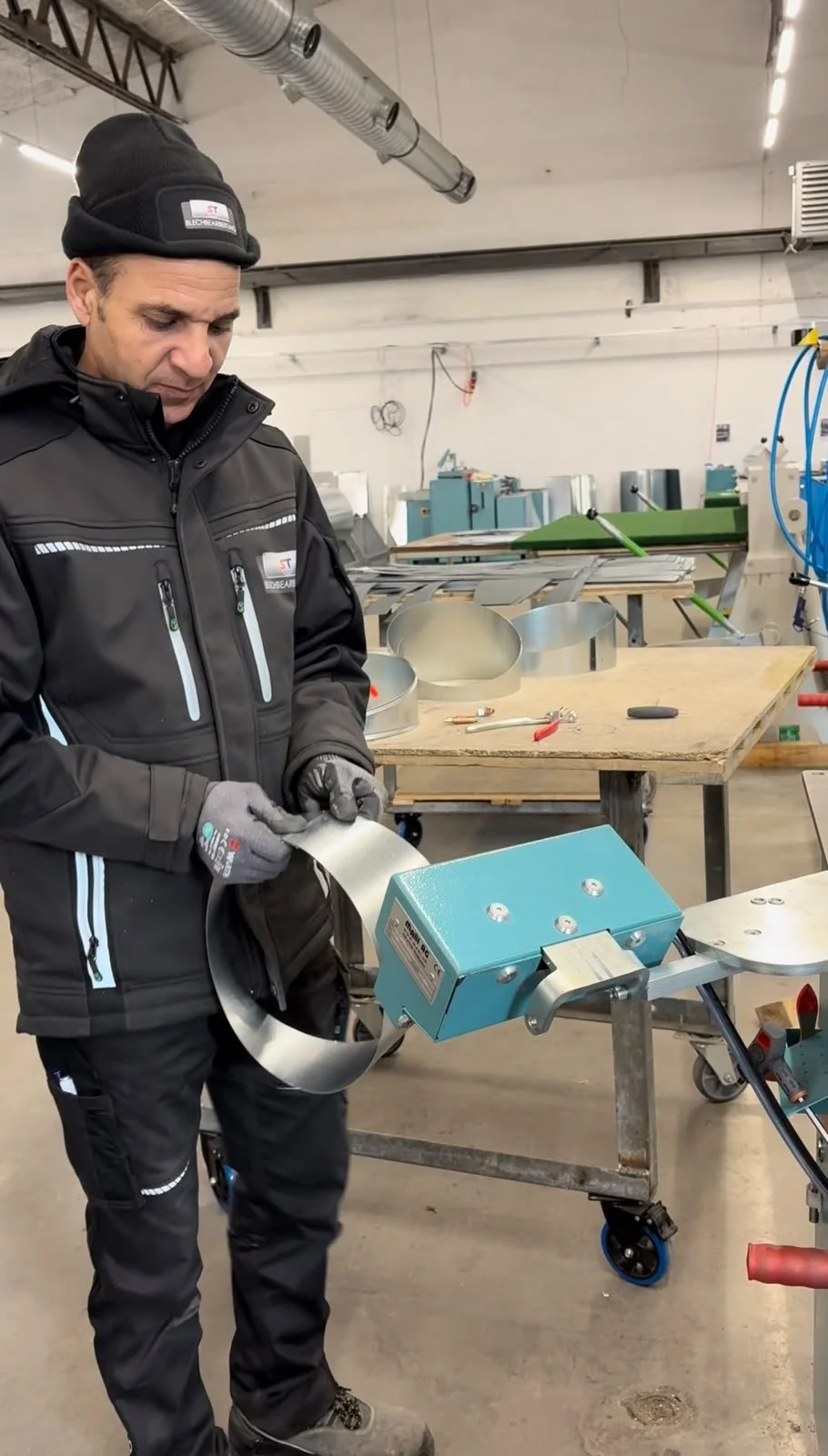 Ein Mann in Arbeitskleidung und Handschuhen hält ein Metallring in einer Werkstatt. Im Hintergrund sind Werkbänke und Maschinen sichtbar.