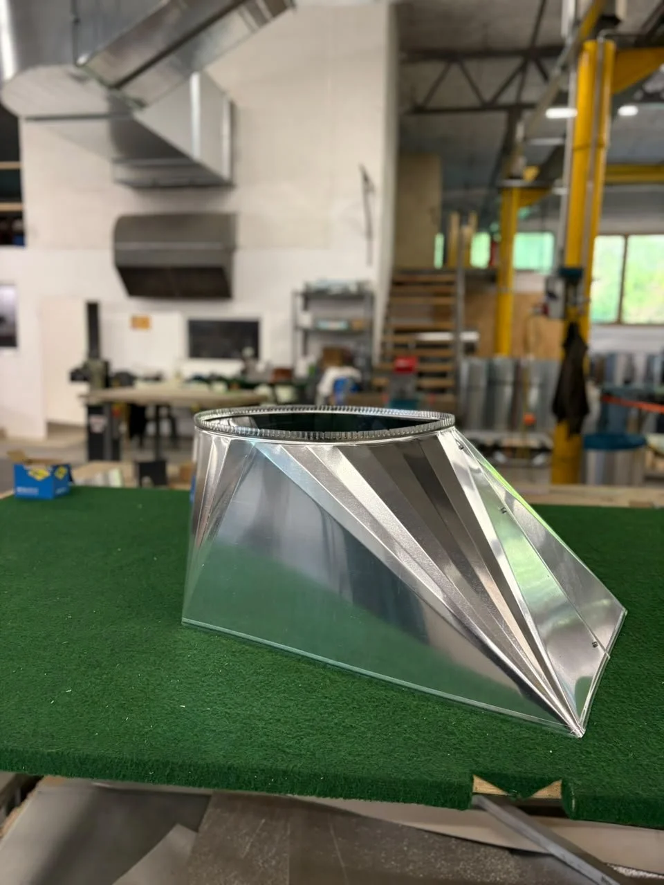 Ein metallischer, sackförmiger Behälter auf einem grünen Tisch in einer Werkstatt, im Hintergrund sind Werkzeuge und Maschinen sichtbar.