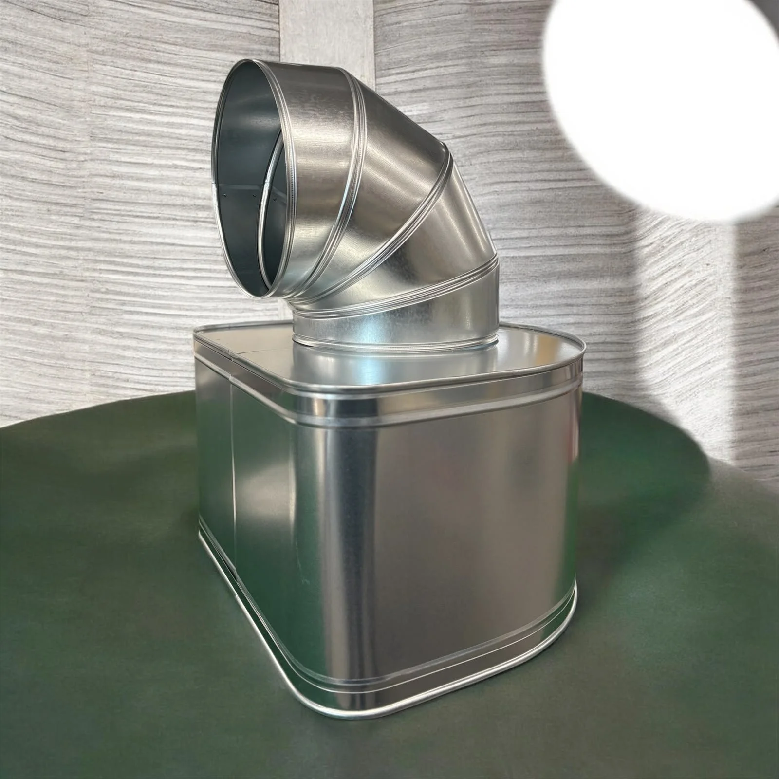 Runde, verspiegelte Metallröhre, die zu einem trichterförmigen Auslass geformt ist, auf einer rechteckigen Metallbasis auf einem grünen Tisch vor einer Wand mit grauer Holzmaserung.