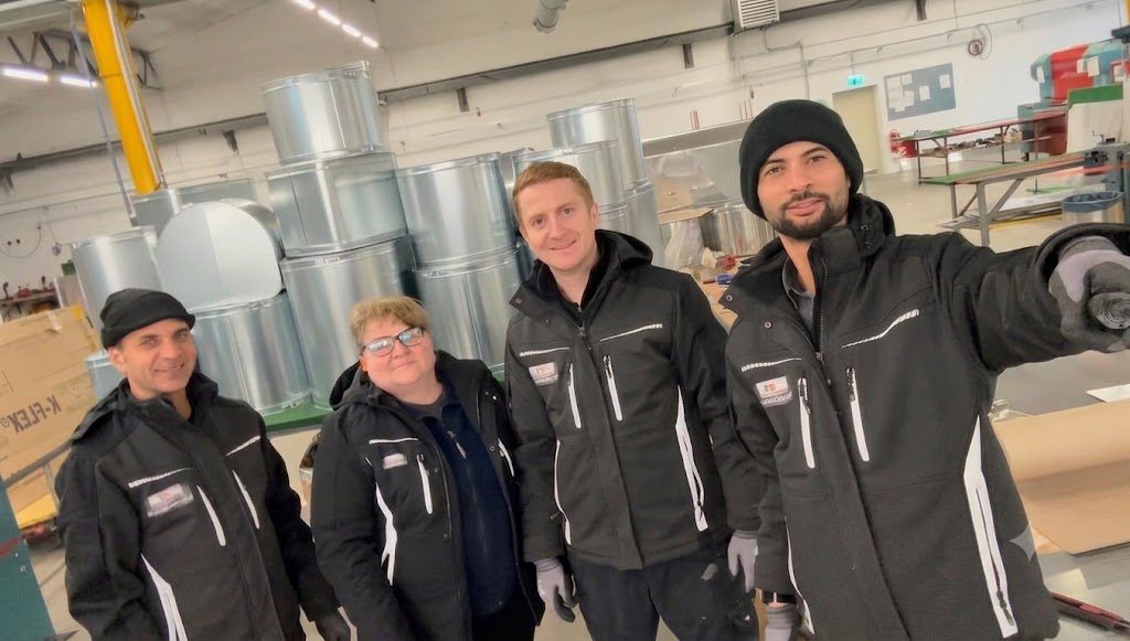 Vier Menschen in schwarzer Arbeitskleidung und Mützen in einer Fabrikhalle, umgeben von Metallrohren und Verpackungsmaterialien.