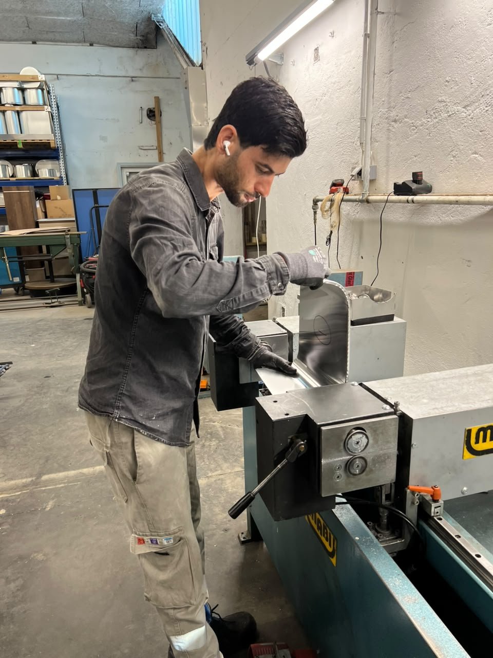Ein Mann arbeitet in einer Werkstatt an einer Metalldrehmaschine, trägt schwarze Handschuhe und AirPods, mit Werkbank und Werkzeugen im Hintergrund.