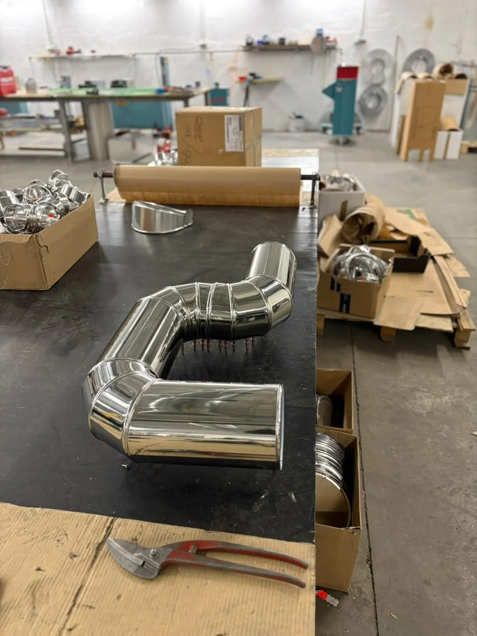 Metallrohre und -teile in einer Werkstatt, möglicherweise für die Chimney- oder Lüftungssysteme, mit Werkzeuge und Verpackungsmaterial im Hintergrund.