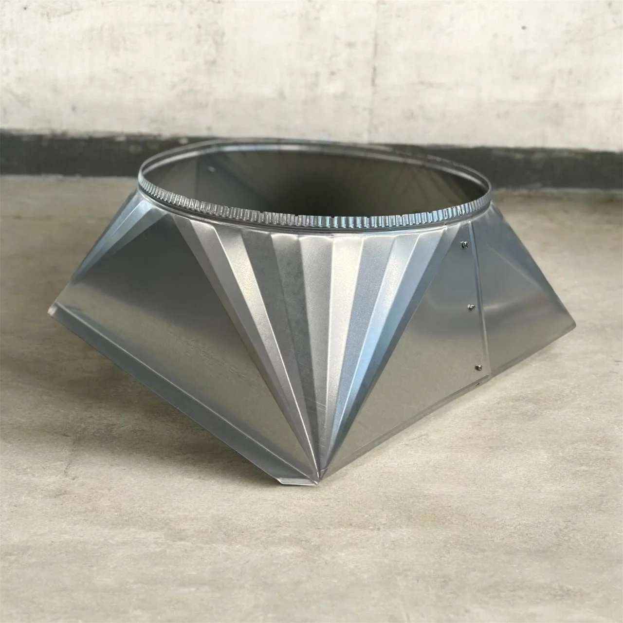 Ein silberner, geometrischer Metallkamin, der wahrscheinlich für einen Kamin verwendet wird, liegt auf einer Holzbodenfläche gegen eine weiße Wand.