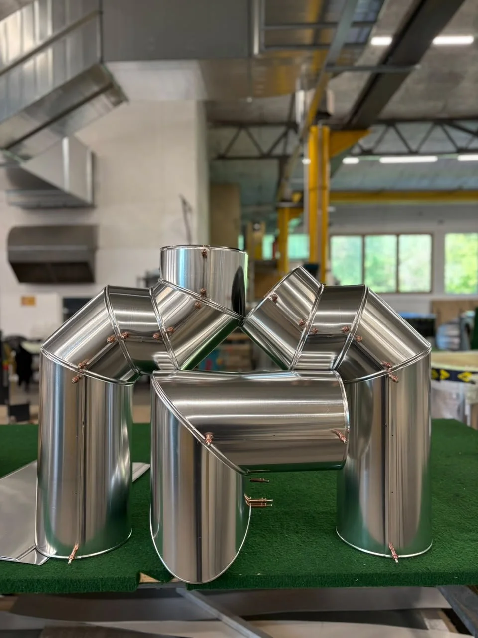 Metallskulptur aus zylindrischen und rechteckigen Rohren, stehend auf einer grünen Oberfläche in einem Industriegebäude.