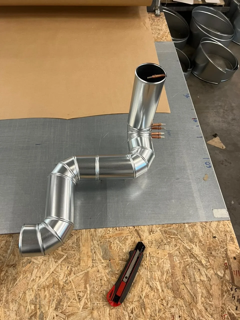 Metallrohrleitung mit mehreren Verbindungsstücken und Schrauben auf einem Werkbank, im Hintergrund Metalltöpfe.