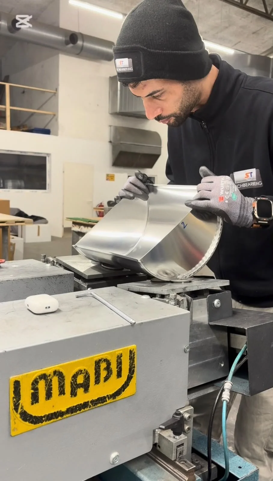 Ein Mann trägt schwarze Kleidung, eine Mütze und Handschuhe, während er ein Aluminium-Teil auf einer Maschine bearbeitet. Der Hintergrund zeigt eine Werkstatt mit Industriegeräten.