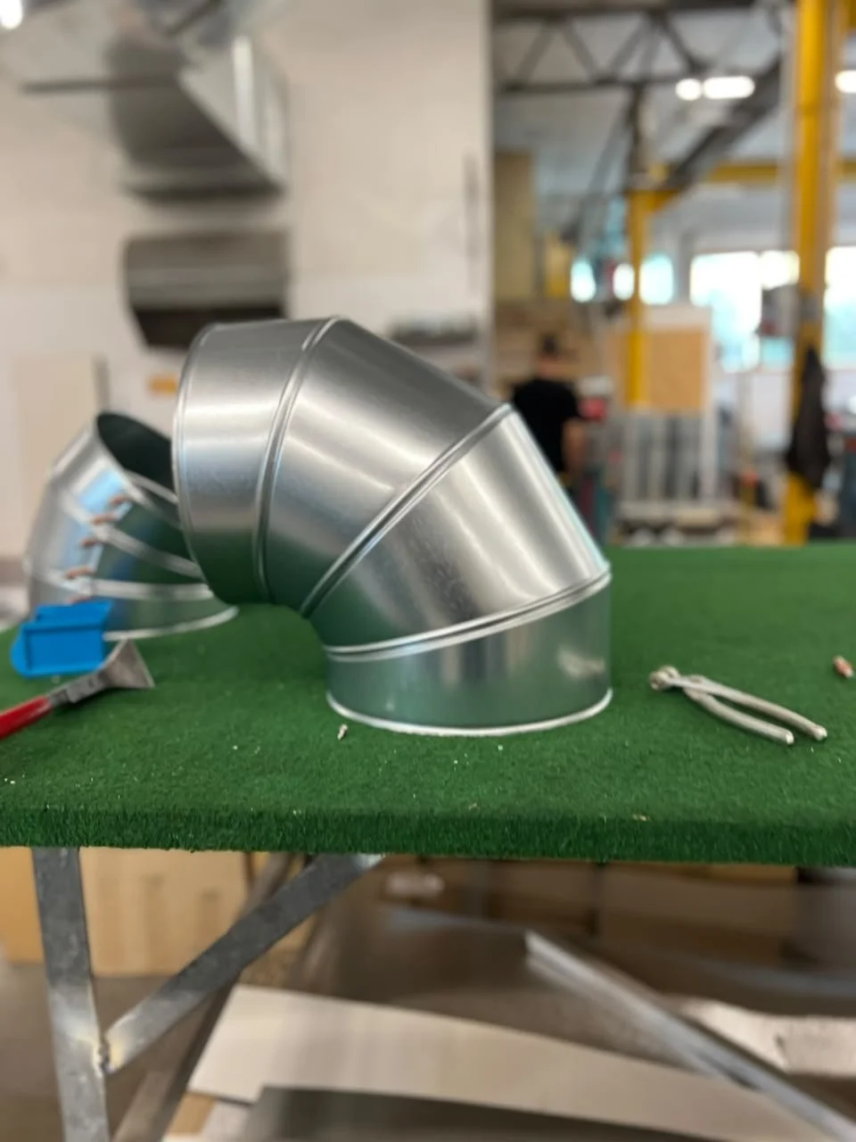 Verzinkte Metallrohre und -stücke, die zu einem Bogen verbunden sind, liegen auf einer grünen Arbeitsmatte in einer Werkstatt.