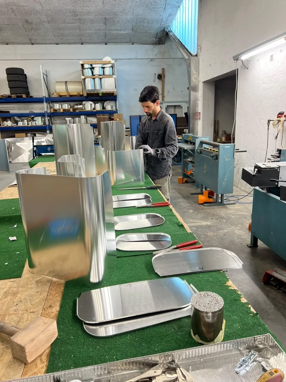 Ein Mann arbeitet in einer Werkstatt an einer Reihe von Edelstahlteilen, die auf einem grünen Arbeitstisch liegen. Es sind verschiedene Metallformstücke zu sehen, einige sind bereits gebogen, andere noch im Rohzustand. Im Hintergrund sind Werkzeuge u