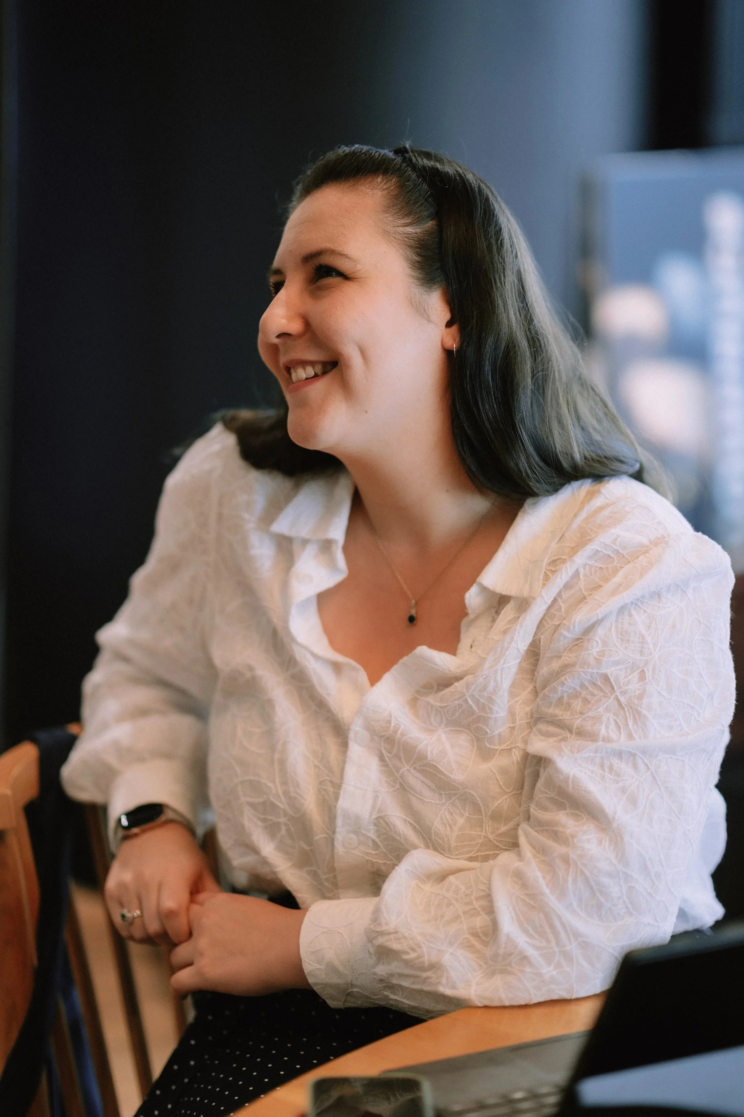 Grace Occleshaw smiling, sitting at a table with her arms crossed. She has dark, shoulder-length hair and is wearing a white patterned blouse, a necklace, and a smartwatch. Grace Thinks about her journey with fondness