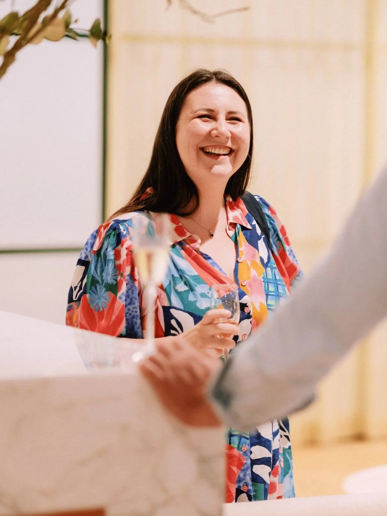 Grace Occleshaw with long dark hair smiling and holding a glass of champagne, wearing a colourful floral dress, at a corporate event. Conversing deeply with an engineer about their business.