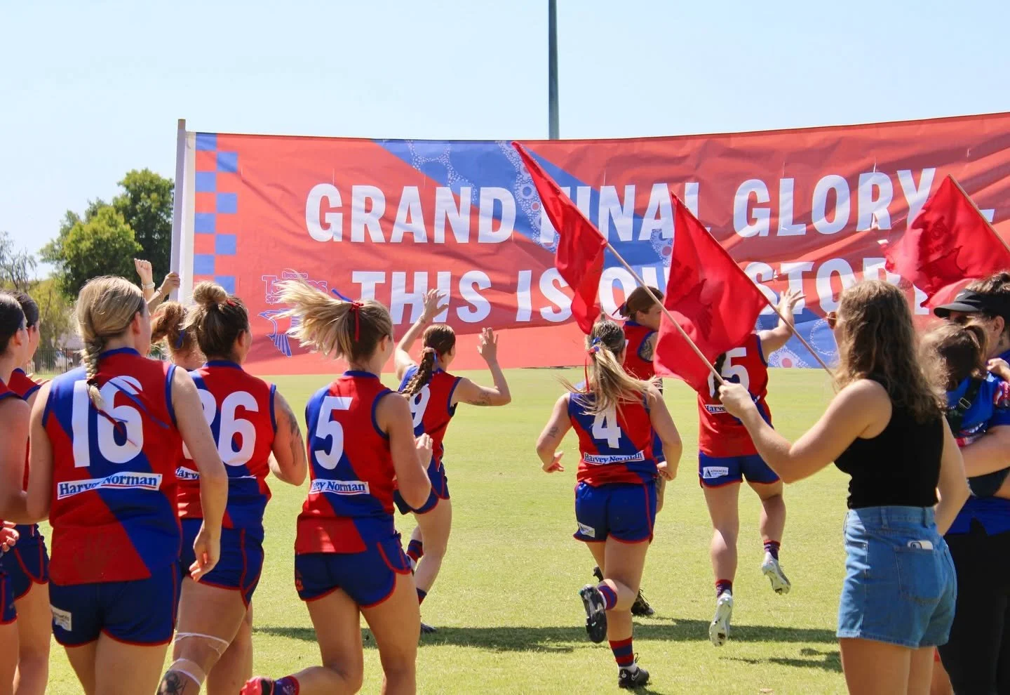 Women athletes in red and blue sports uniforms running onto a field with a large banner that reads 'Grand Final Glory, This is Our Story' and holding red flags.