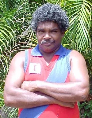A man with curly black hair and a mustache, wearing a blue and red sleeveless shirt, standing outdoors in front of green tropical plants with crossed arms.