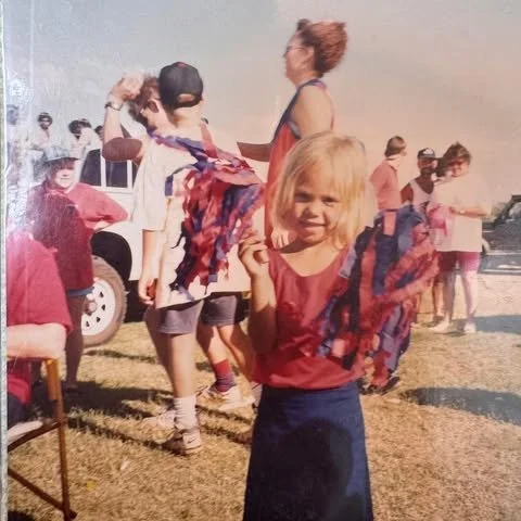 A group of children and adults gathered outdoors. A young girl with blonde hair in a red shirt and dark skirt stands in the foreground, smiling and holding a patterned scarf. Other children and adults are seen in the background near parked vehicles, with some children playing and others standing around.