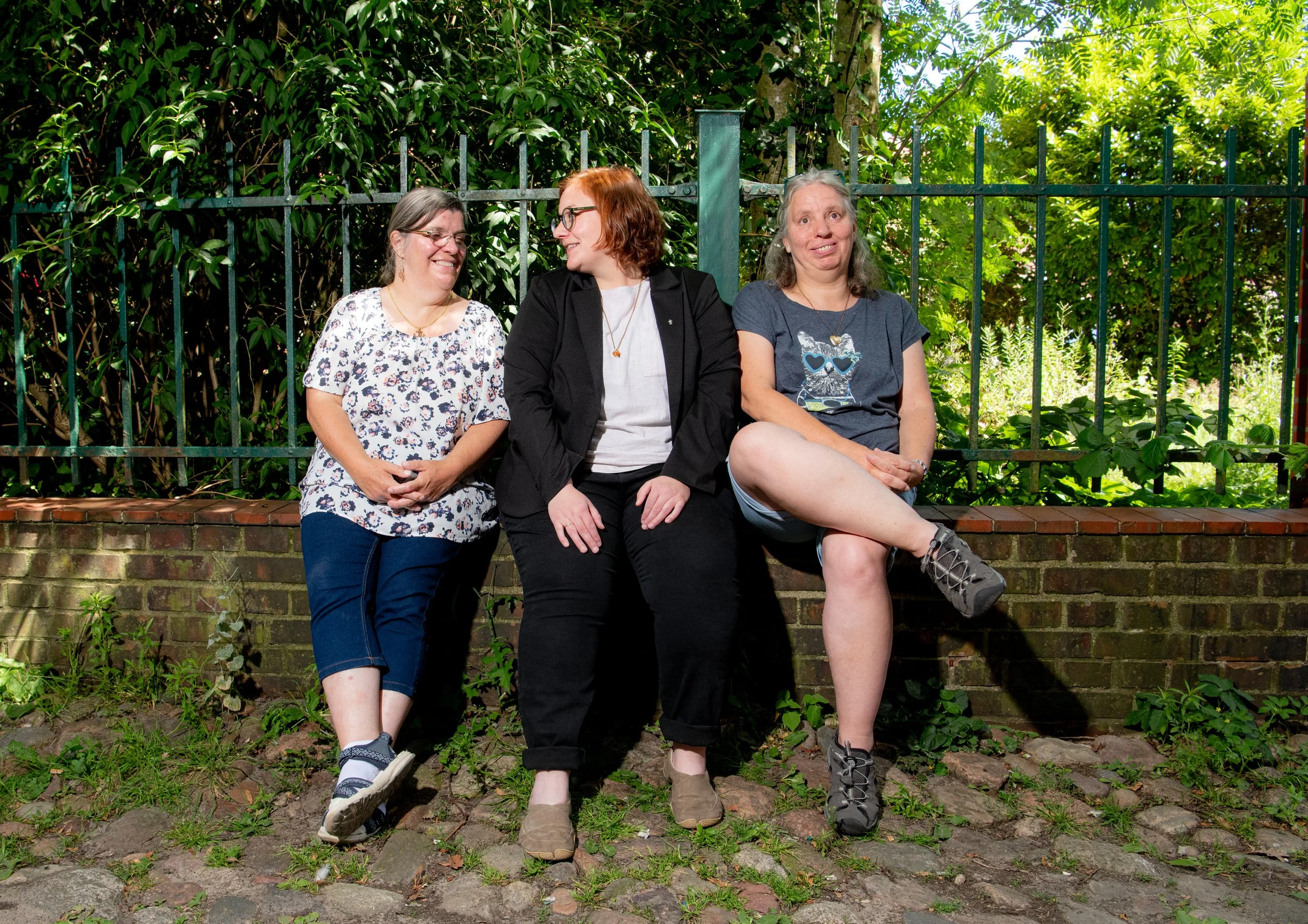 Drei Frauen sitzen nebeneinander auf einer Mauer im Freien, umgeben von grünen Pflanzen und Bäumen, lachen und unterhalten sich.