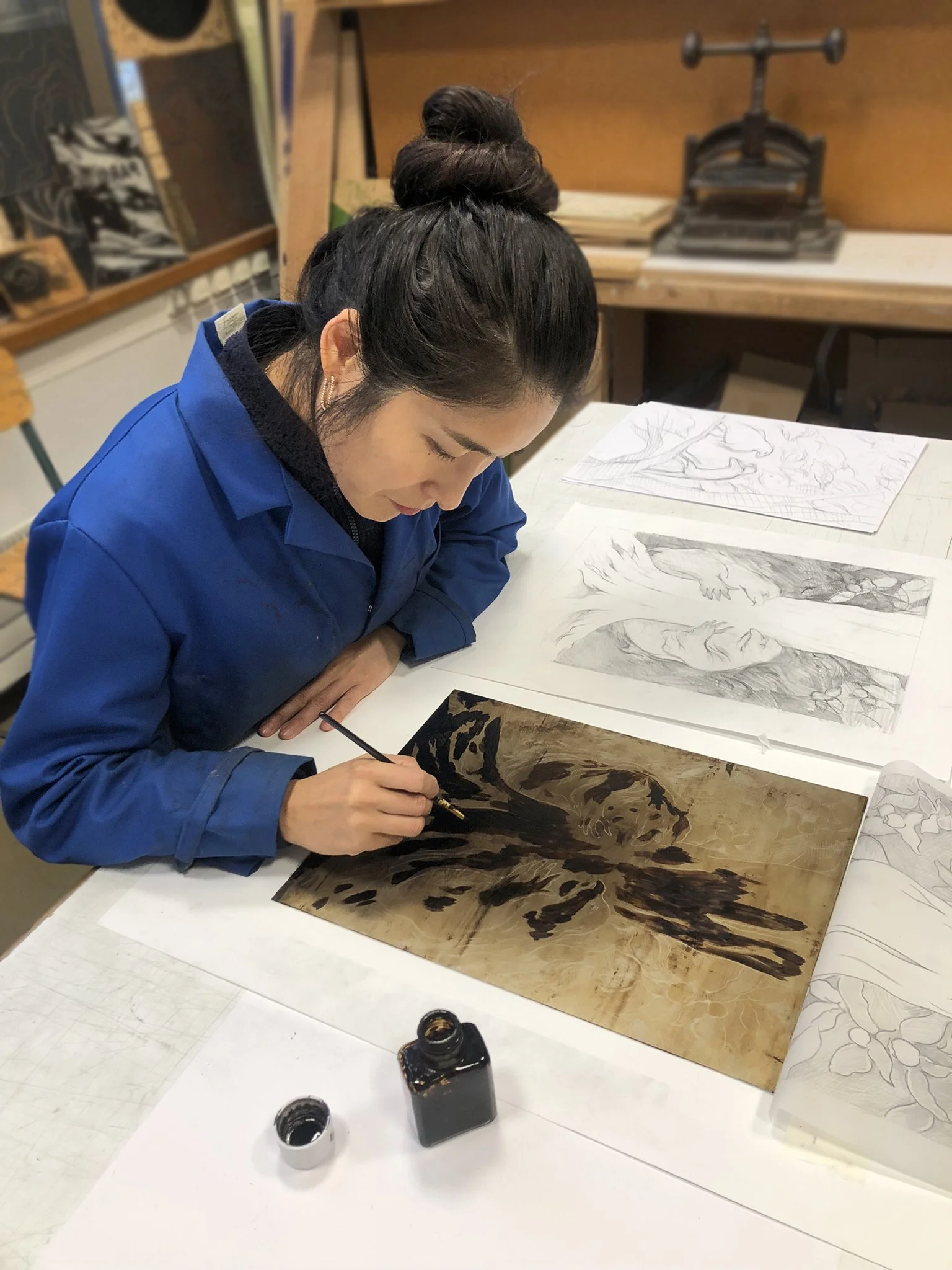 Une femme dessine des croquis de paysages et de nature avec un stylo à un bureau, entourée de livres et d'encre.