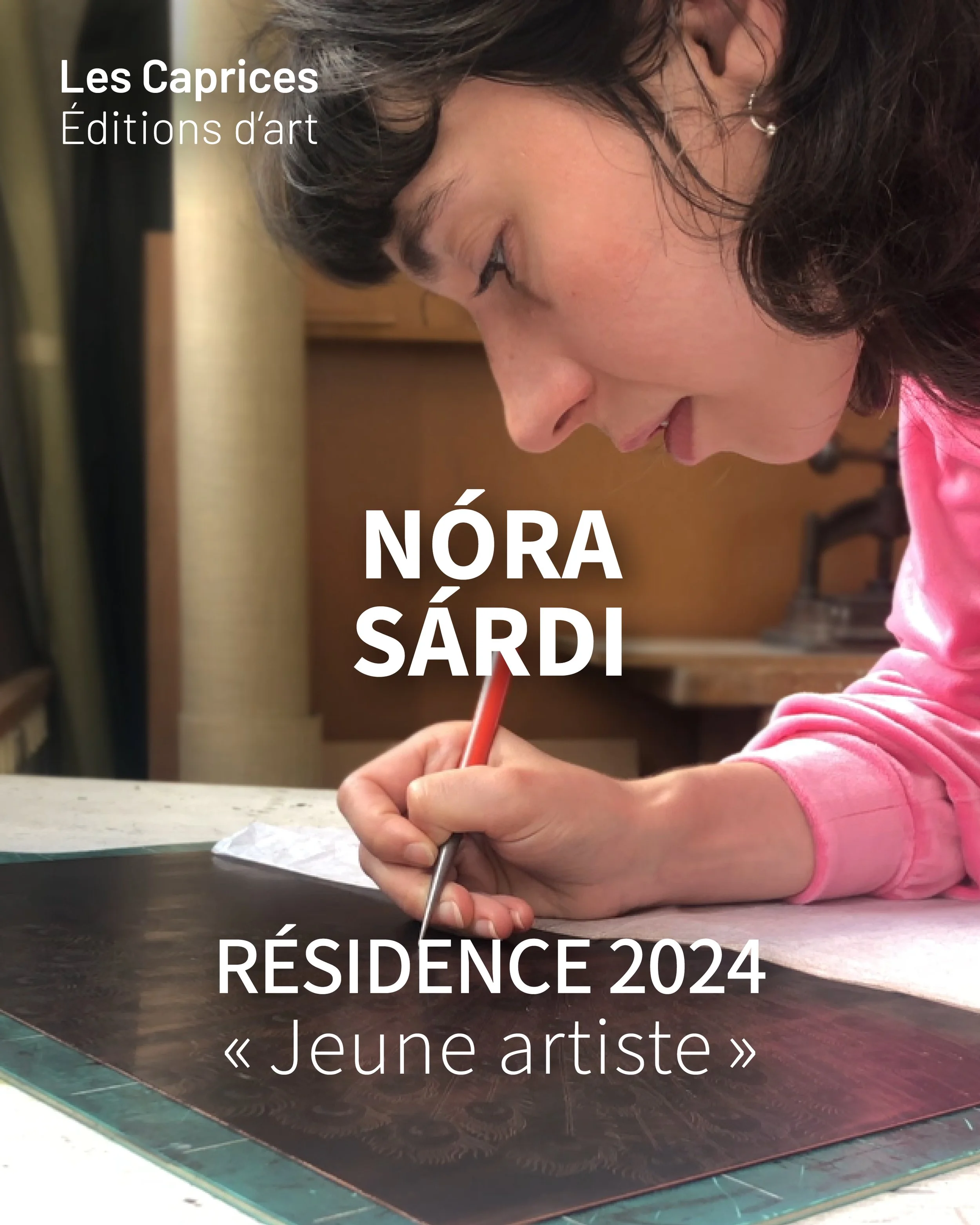 Une fille écrivant avec un crayon sur une table en bois clair, à l'arrière-plan une étagère en bois et un mur beige. La fille porte un sweat rose et une boucle d'oreille en perle.