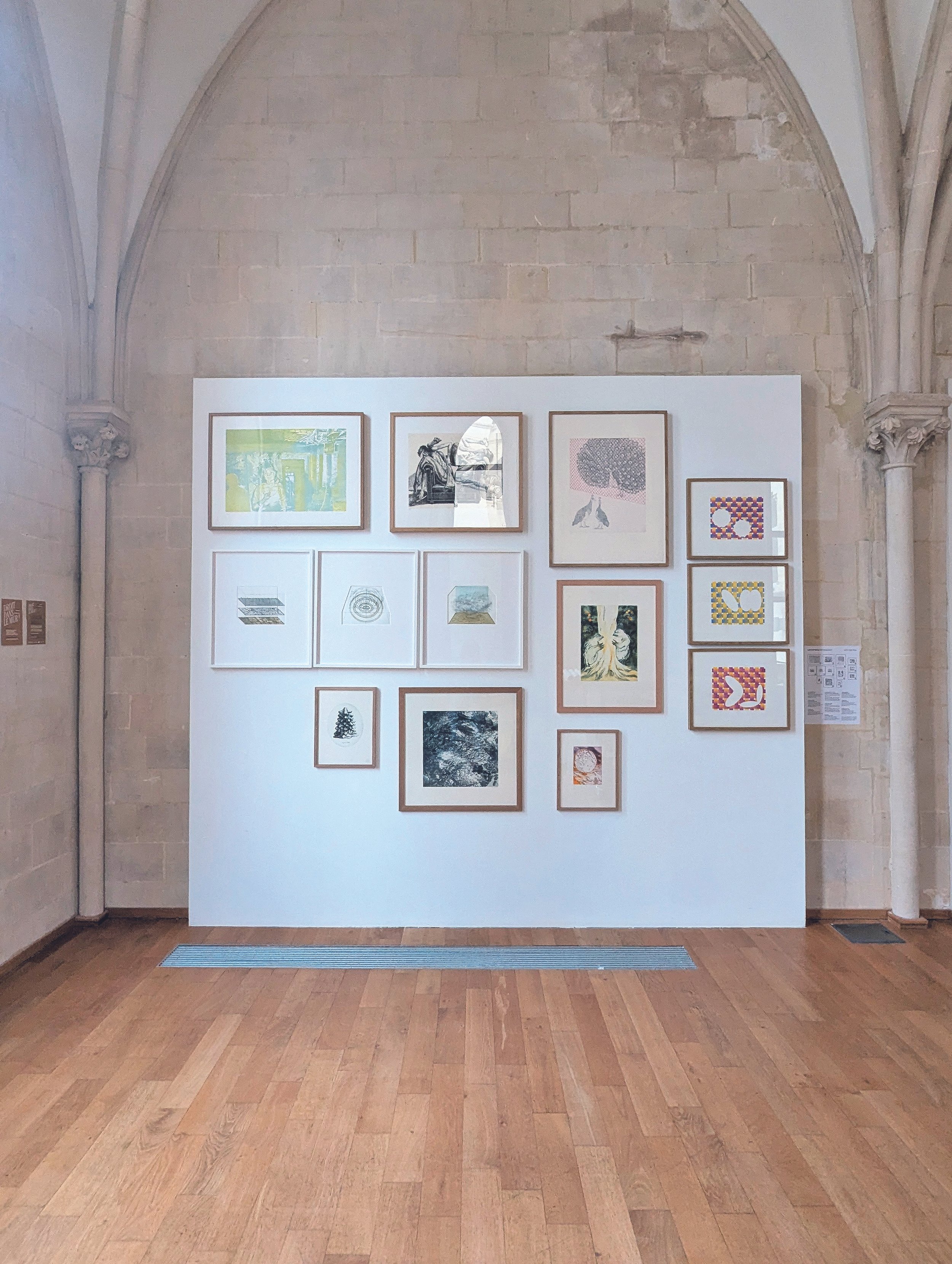 Exposition d'art avec plusieurs œuvres encadrées accrochées sur un mur blanc, dans une salle avec murs en pierre et sol en parquet.