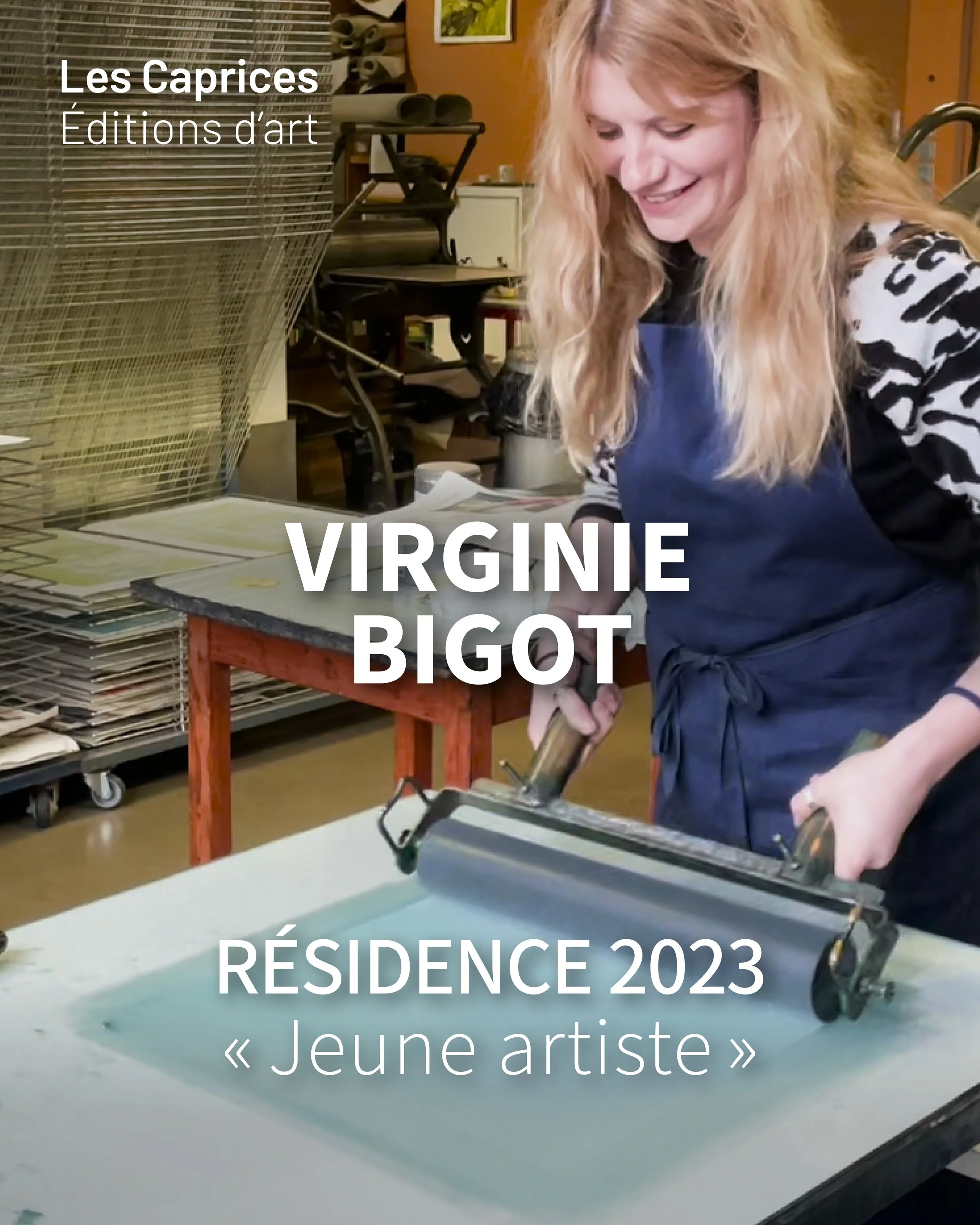Une femme aux cheveux longs et blonds, portant un tablier bleu, travaille dans un atelier, en train de scier du contre-plaqué. Sur la gauche, une étagère avec des rouleaux de papier ou de toile, et derrière elle, il y a une table de travail. L'atelier a des murs en bois, avec des affiches ou des images accrochées. L'image est utilisée pour un article sur une jeune artiste, Virginie Bigot, pour une résidence en 2023.
