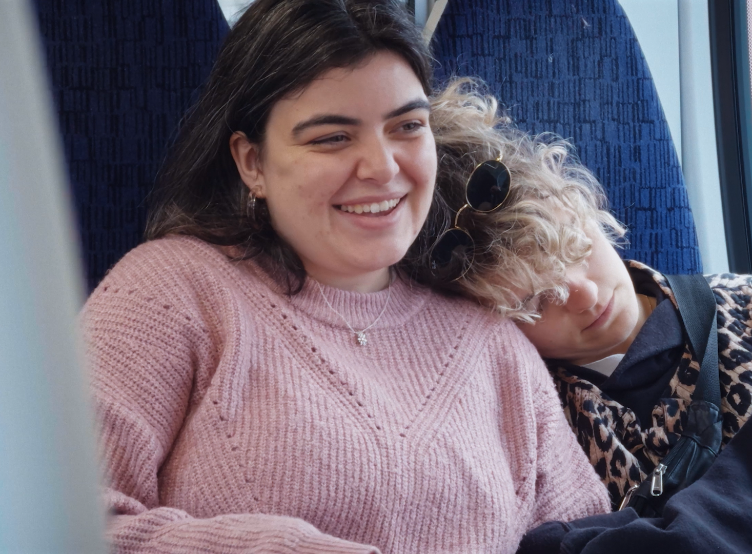 A young woman with dark hair smiling while sitting next to a young boy with curly blond hair resting his head on her shoulder on a train.