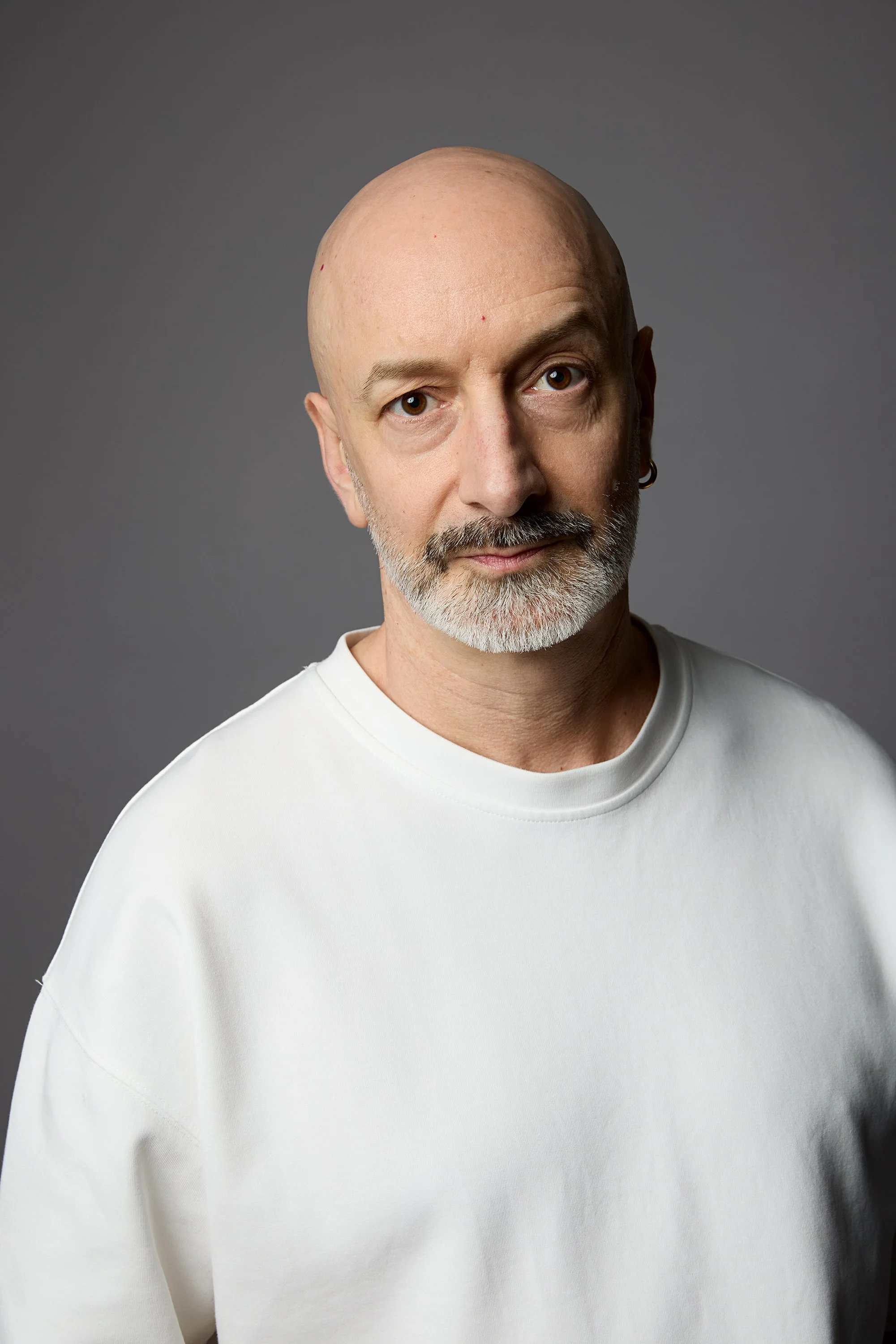 A middle-aged man with a bald head and gray beard wearing a white shirt, standing against a plain gray background.