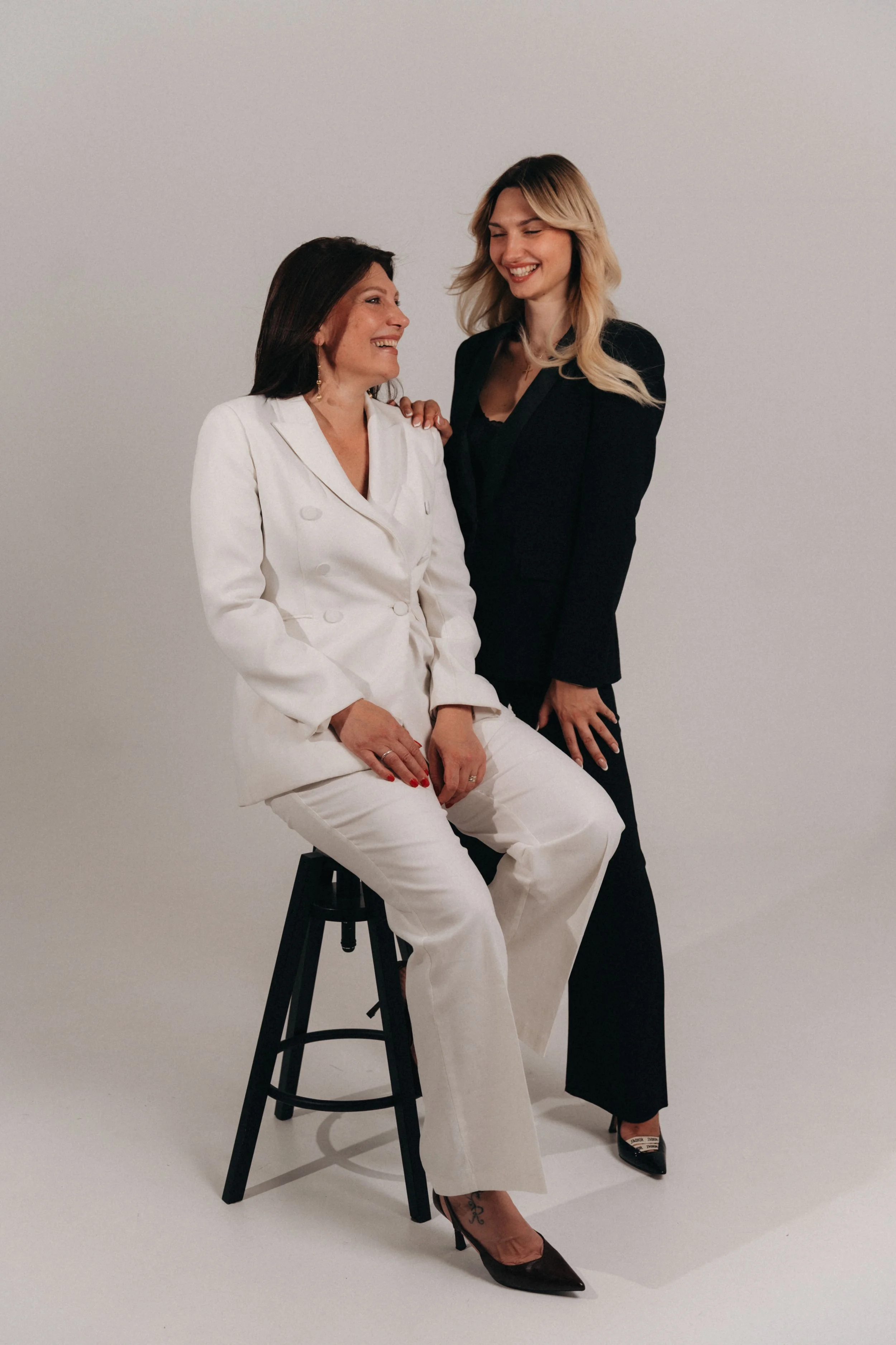 Due donne sorridenti, una seduta su uno sgabello nero con abito bianco e l'altra in piedi con abito nero, in uno studio con sfondo neutro.