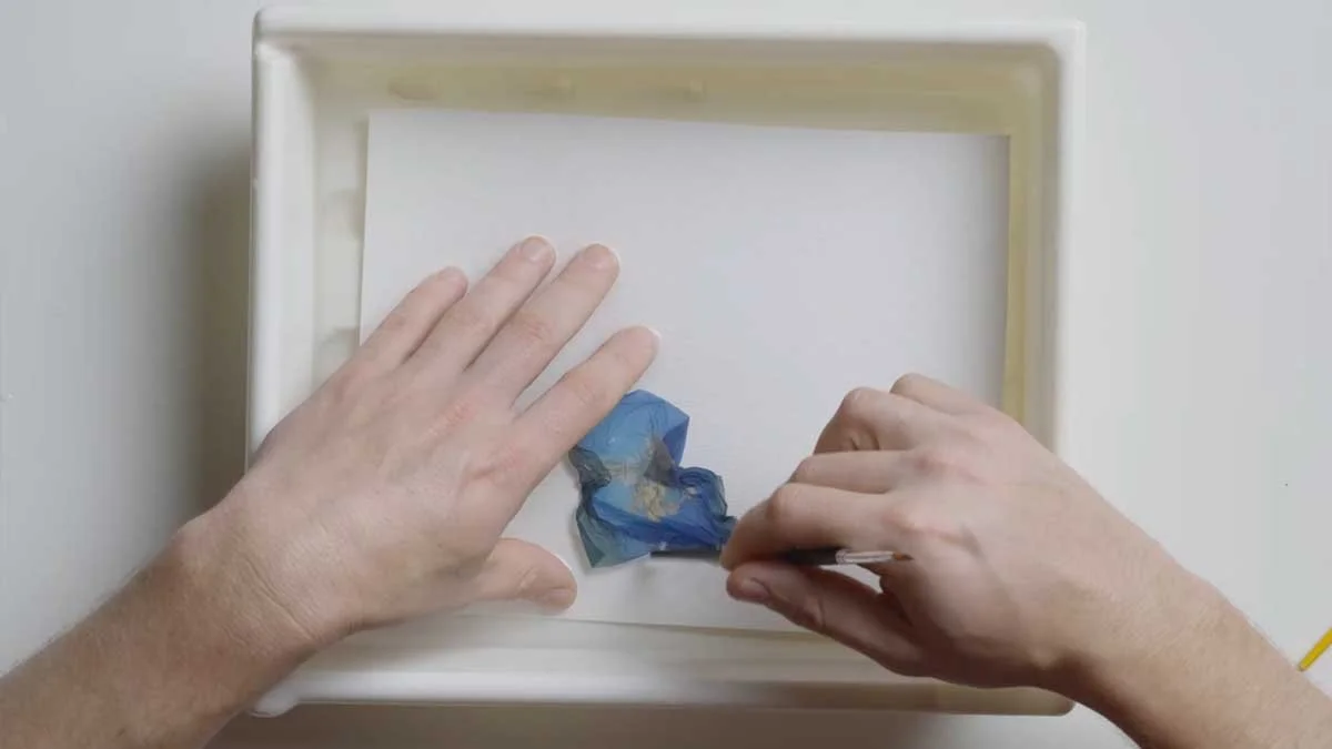 A pair of hands moving the emulsion around over the top of a piece of paper.