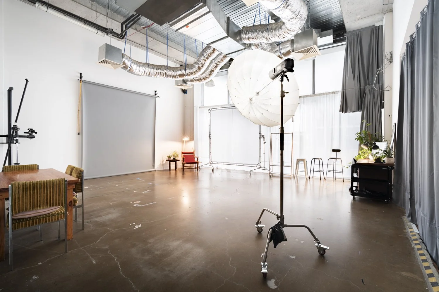 A large studio with high ceilings filled with natural light. There is a roller light stand with a godox light and a shoot through umbrella. There is a grey paper backdrop, a large scrim, chairs and curtains which can block out the outside light.