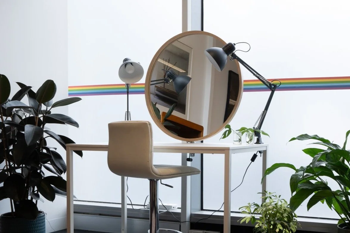 Makeup table positioned beside large frosted windows with natural light, featuring a round mirror, adjustable lamps, a salon chair and indoor plants.