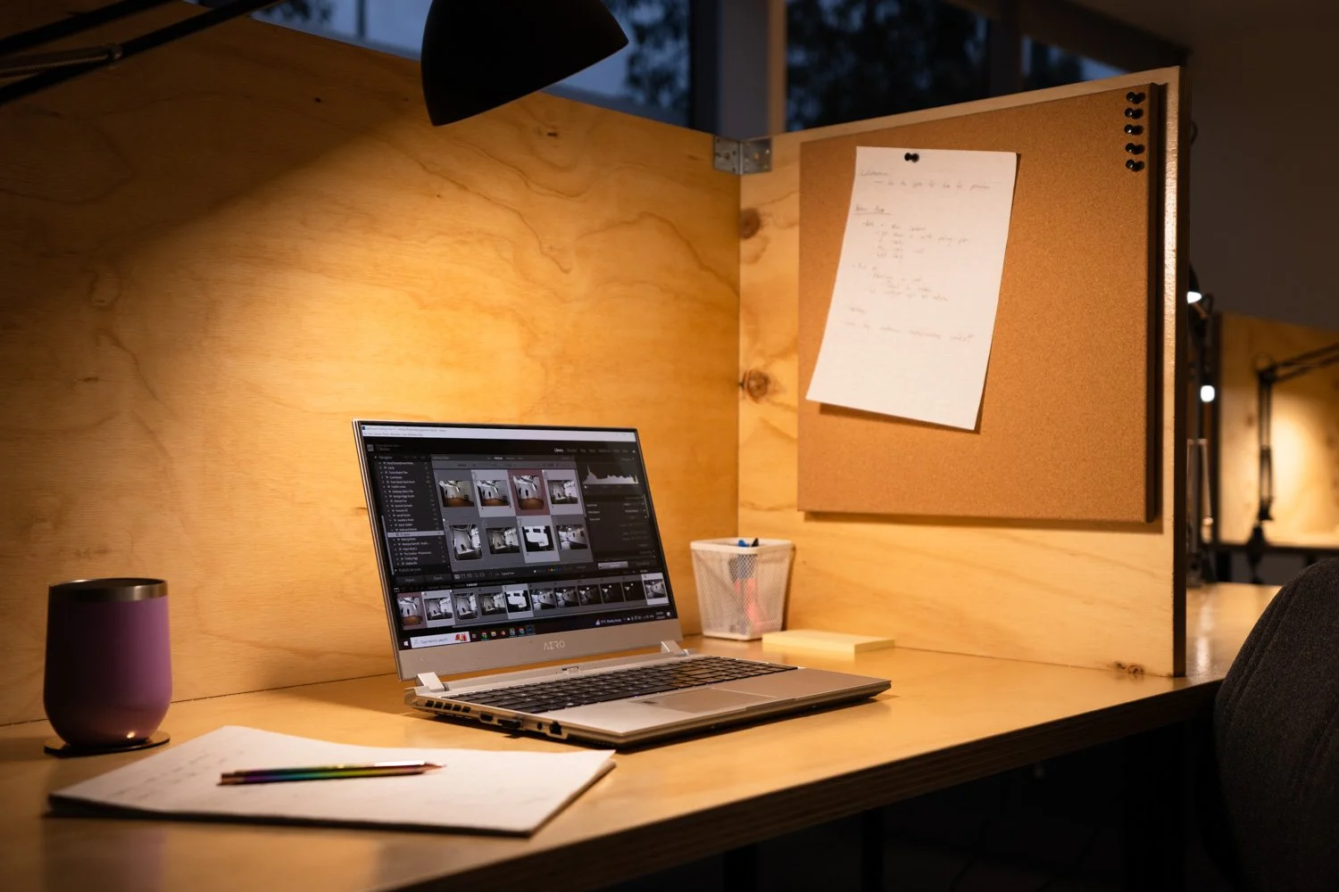 Warm timber workspace with a laptop, desk lamp, notebook, pencils, pinboard and office chair, set up as a quiet editing or admin desk inside a creative studio.