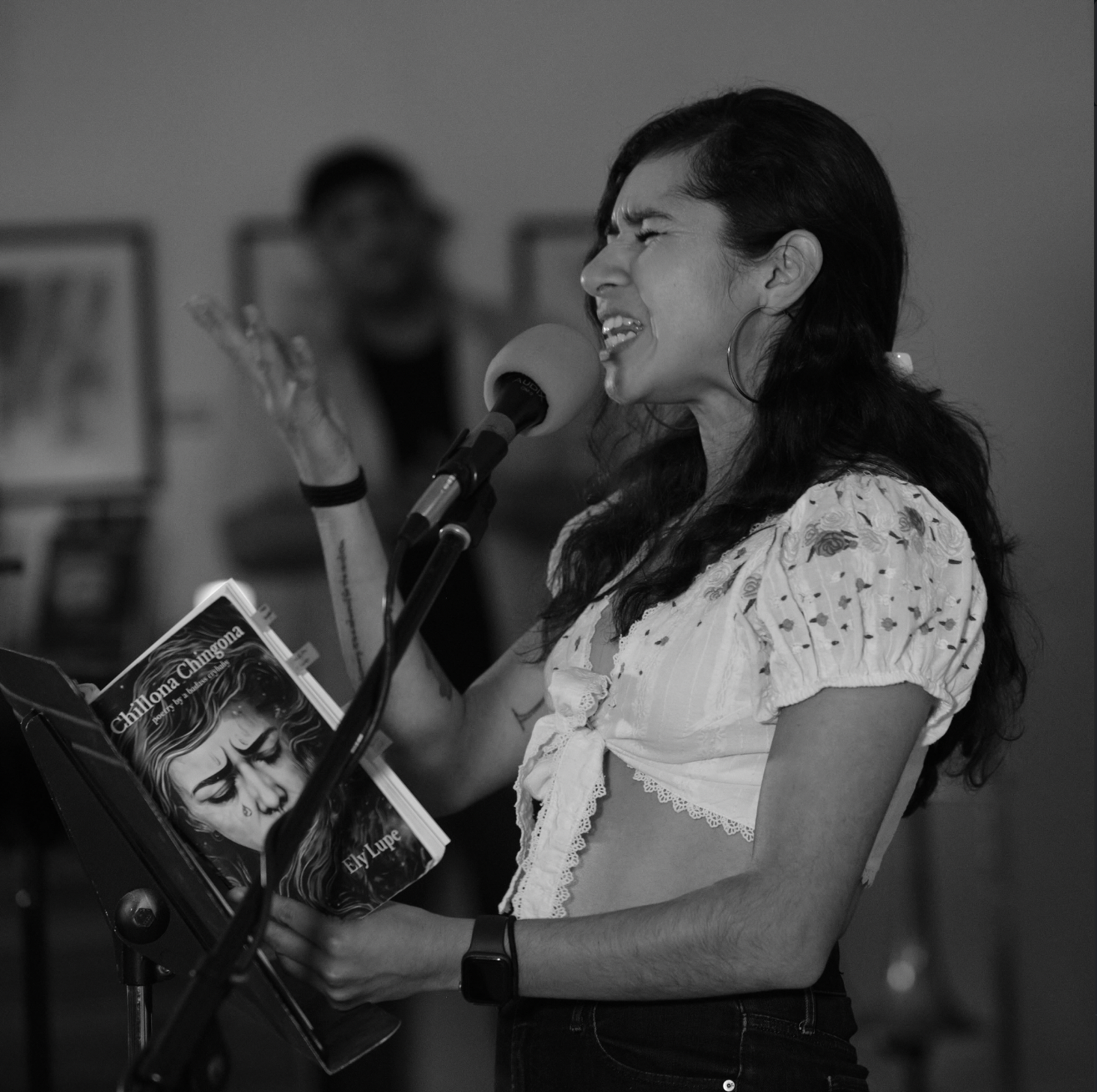 A moment from Ely Lupe reading her poem "The Greatest Country in the World" from her debut book of poetry 'Chilona Chingona' at a May Day Open Mic. She is wearing a white, lacy crop top and large hoop earrings.