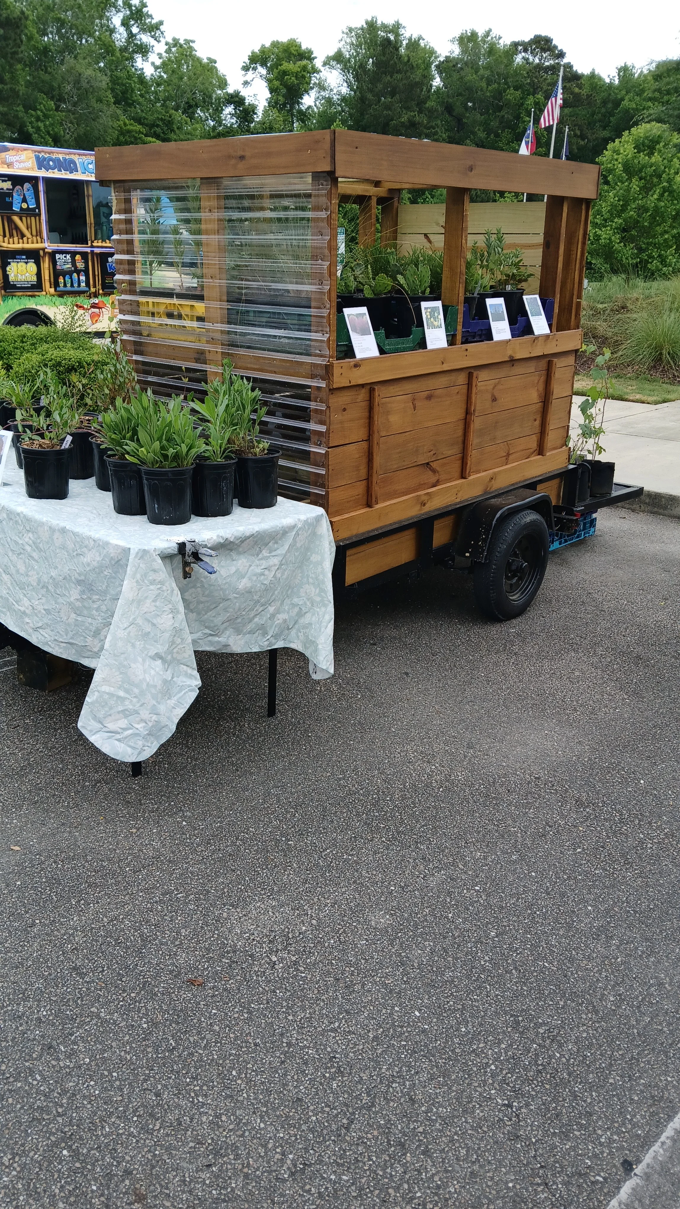 A small mobile plant stand with a wooden structure and clear plastic siding, featuring potted plants. Sells large variety of native plants in spring in Wilmington metro area. See Facebook page for more information