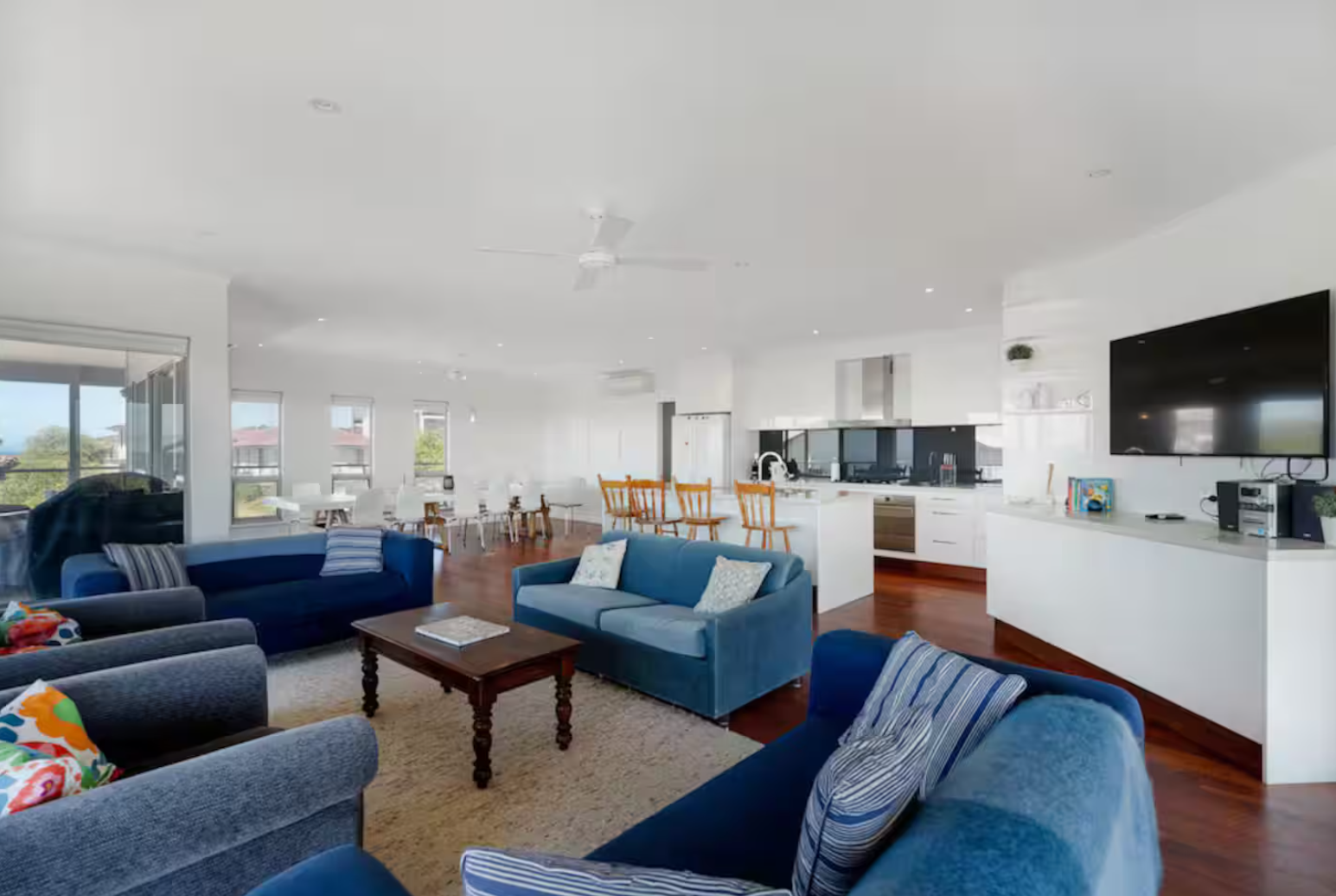 Spacious open-concept living room with blue sofas, coffee table, and a large flat-screen TV on the wall. The room has hardwood floors and large windows letting in natural light. The background shows a kitchen with white cabinets, a breakfast bar with wooden chairs, and a dining area with white chairs.