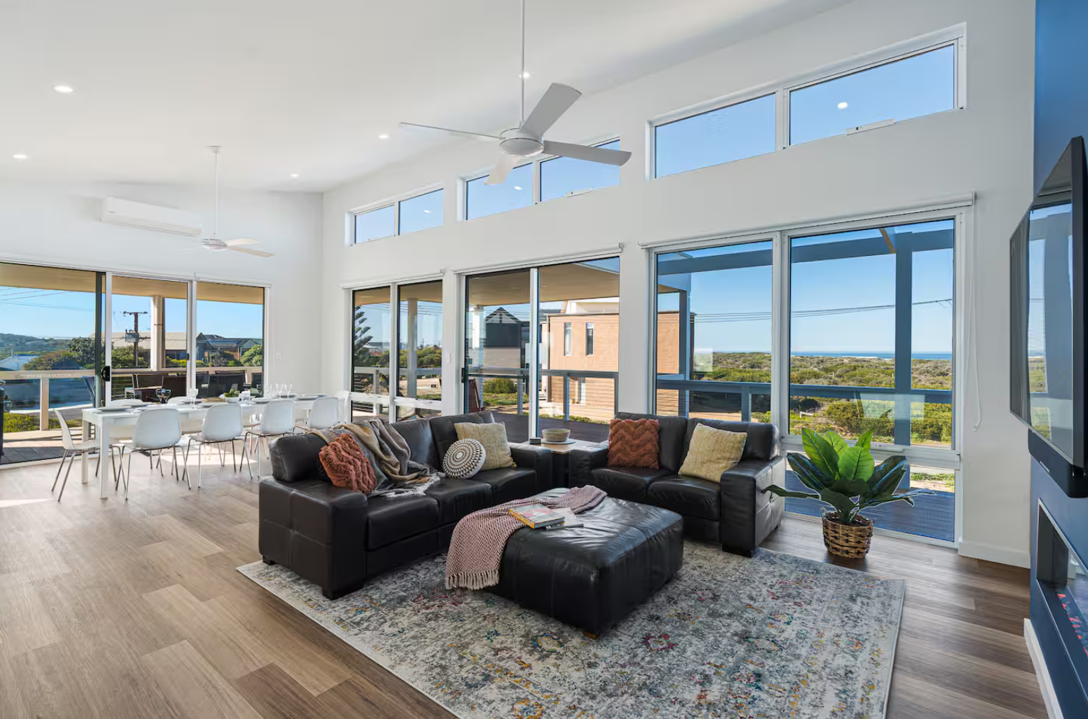 Spacious living room with large windows and glass doors overlooking an outdoor deck and view of greenery and ocean in the distance; modern furniture including a black leather sectional sofa with colorful pillows, a patterned area rug, and a potted plant, with a dining area in the background.