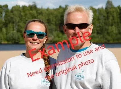 Two smiling people, a woman and a man, wearing sunglasses and white t-shirts with "SWIM LUAND" logo, outdoors near a body of water with green trees in the background.