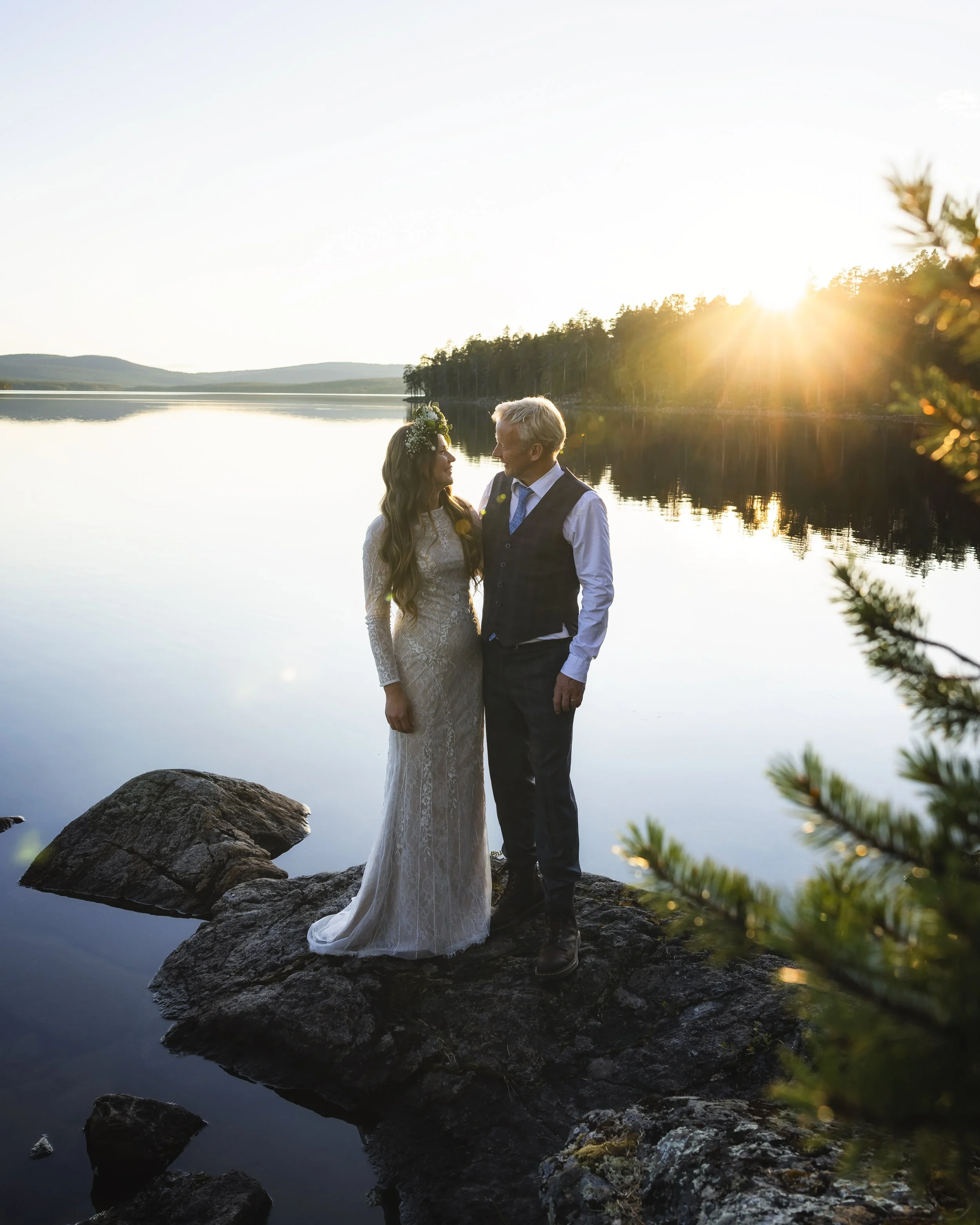 A couple dressed in wedding attire standing on rocks by a calm lake during sunset, with trees and distant hills in the background.