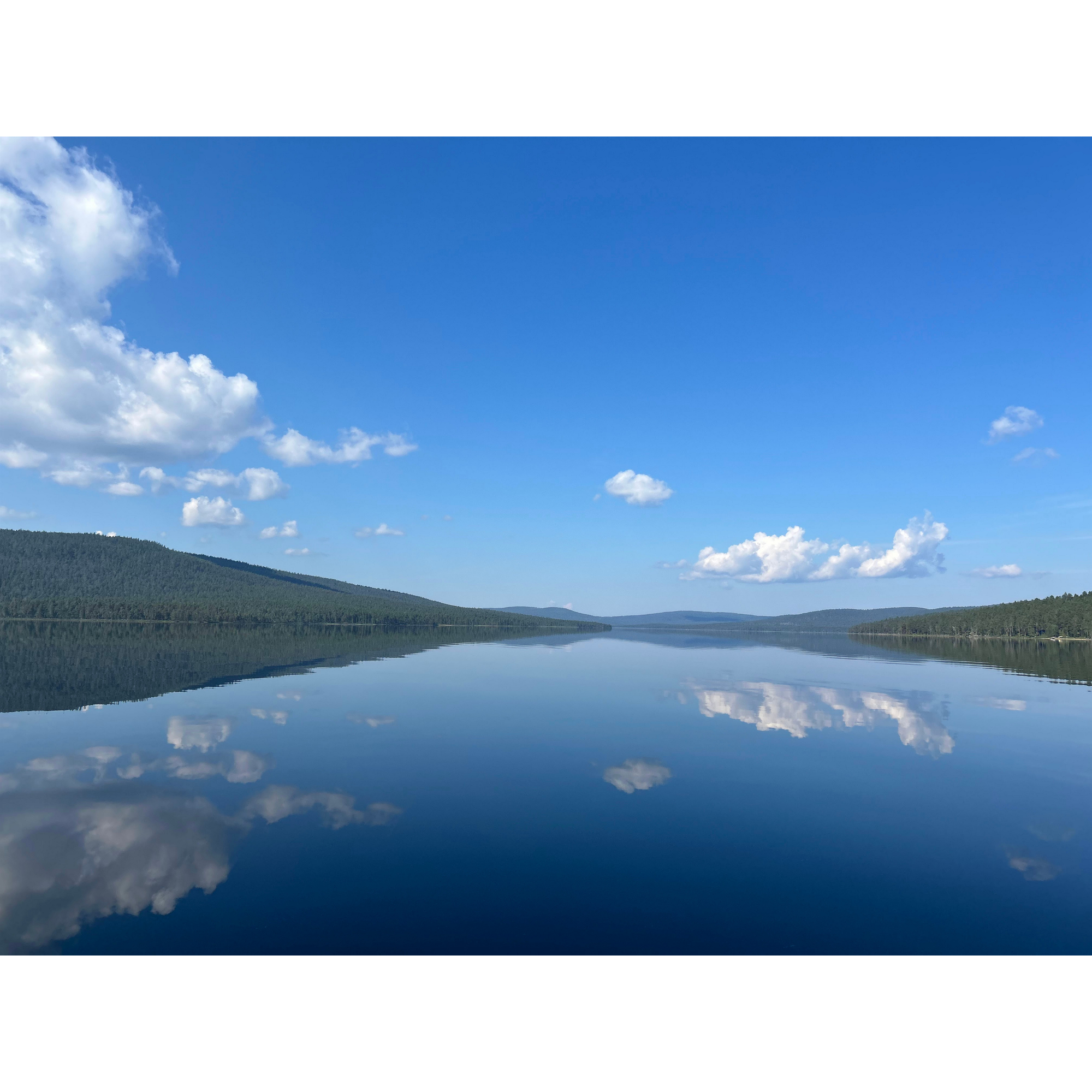 A scenic landscape with a calm lake reflecting the blue sky and white clouds, surrounded by green forested hills.