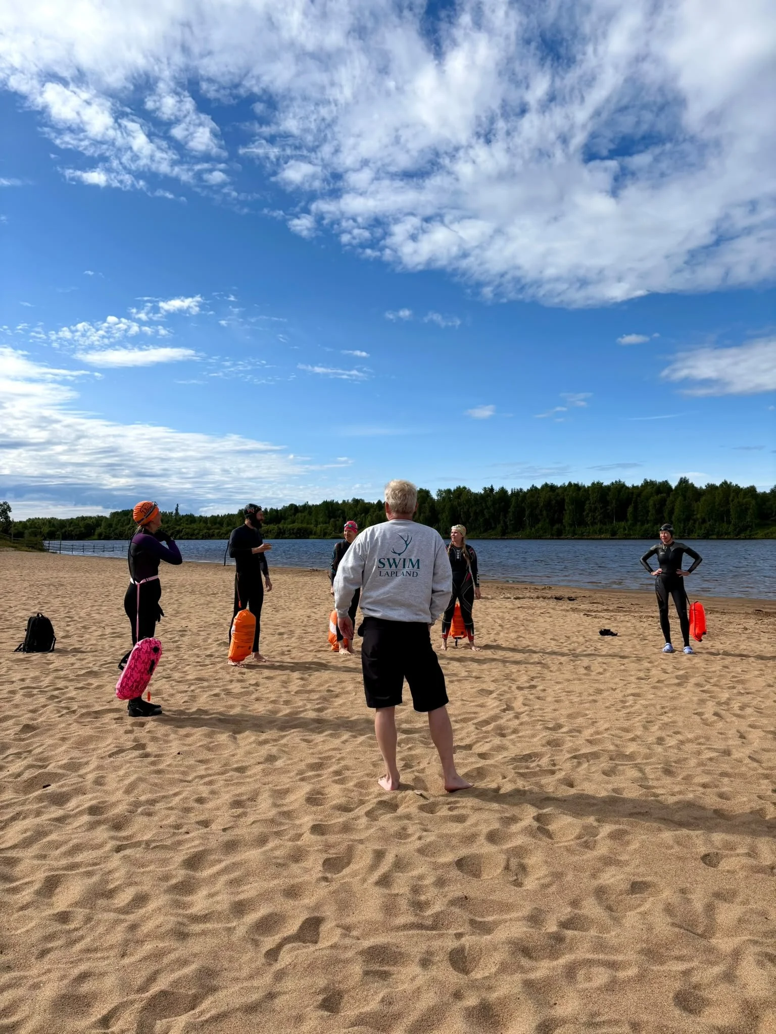 Group of people in swim gear and wetsuits standing on sandy beach near a lake, some holding flotation devices, with trees and a partly cloudy sky in the background.