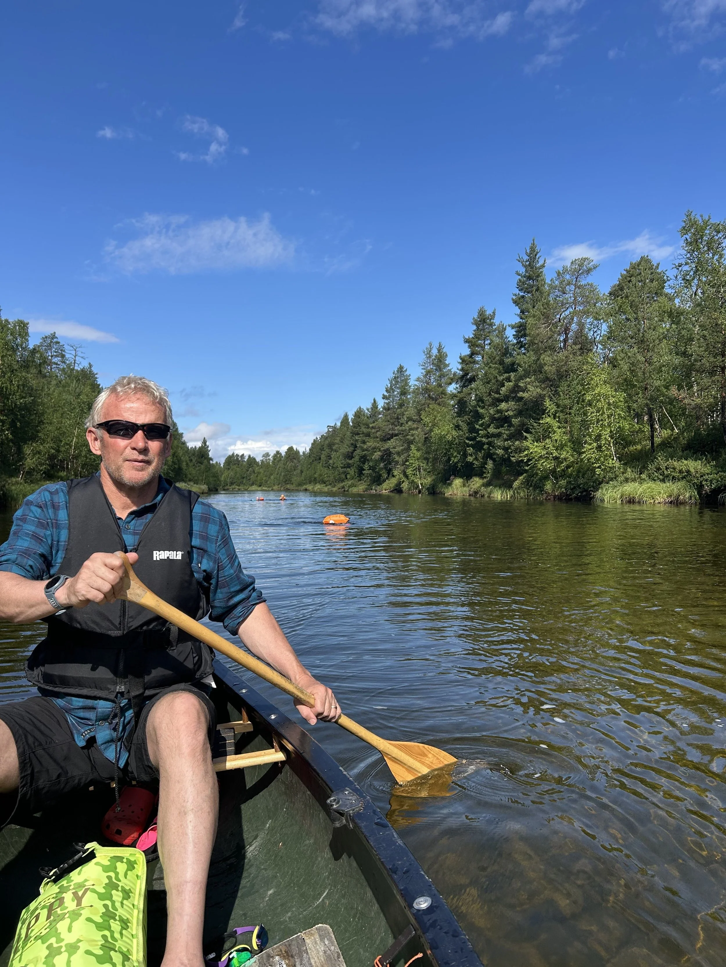 A man wearing sunglasses and a life vest sitting in a canoe on a river, paddling on a bright, sunny day with a clear blue sky and green trees along the riverbank.