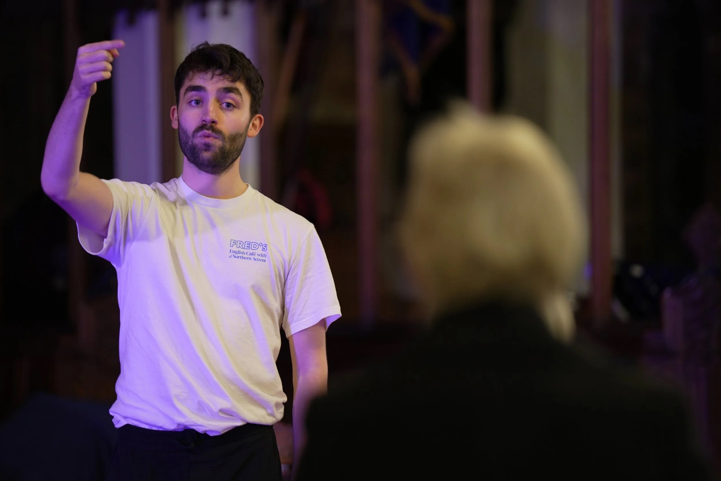 A young man with dark hair and a beard raising his right arm with his hand near his head, standing in a dimly lit room with purple lighting. He is wearing a white T-shirt with text that reads 'FRED'S English Café with a Northern Accent.' He is looking directly at the camera with a slightly pursed expression. There are blurred figures and structures in the background.