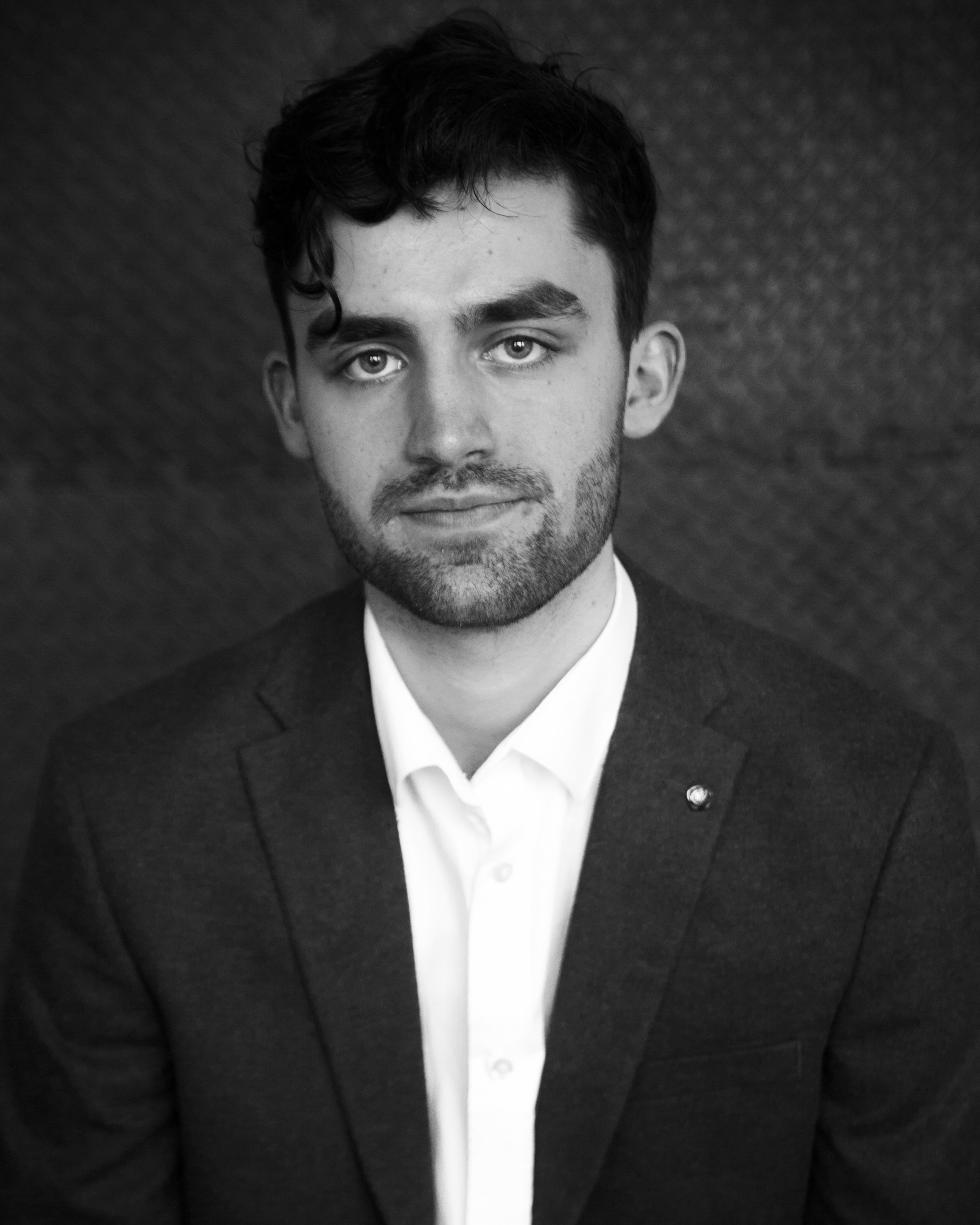 A black and white portrait of a young man with dark, wavy hair, light eyes, and a well-groomed beard, dressed in a dark blazer and a white shirt, against a dark textured background.
